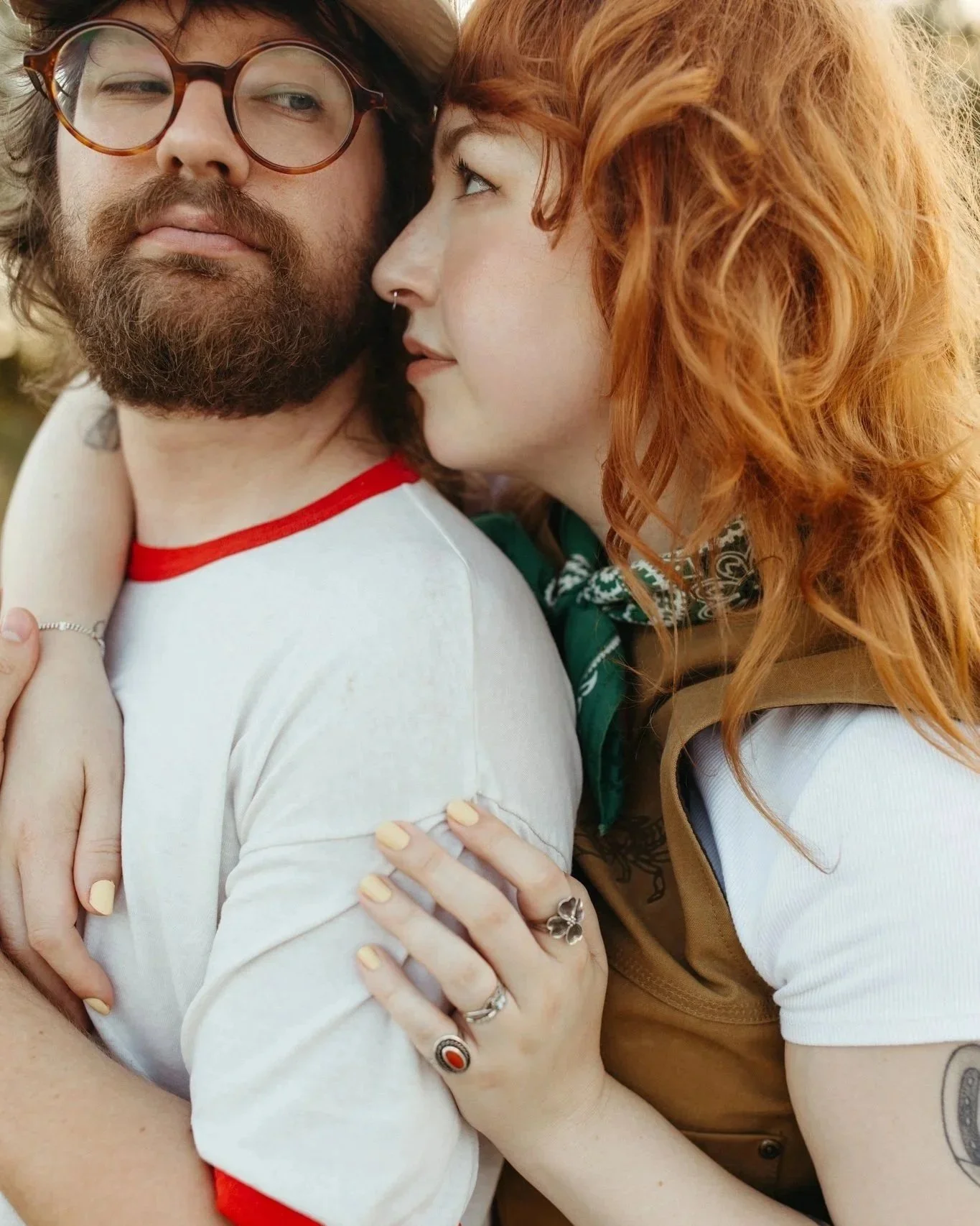 A young woman with curly red hair and fair skin, embracing and close to a bearded man with glasses, brown hair, and fair skin, outdoors.