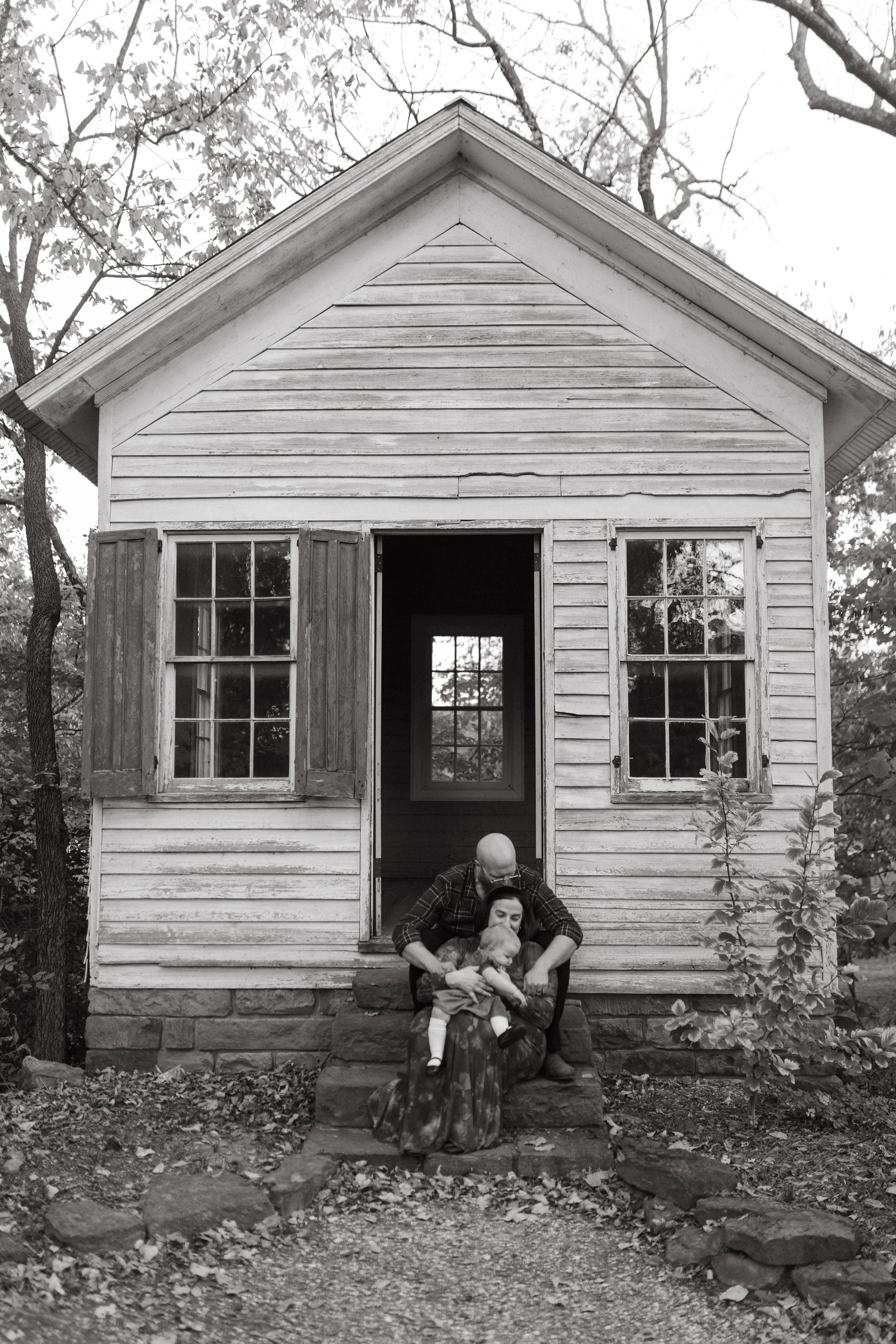 A black and white photo of a small wooden house with an open door and two windows with shutters, surrounded by trees. A person with a bald head and glasses is sitting on the steps holding a young child.