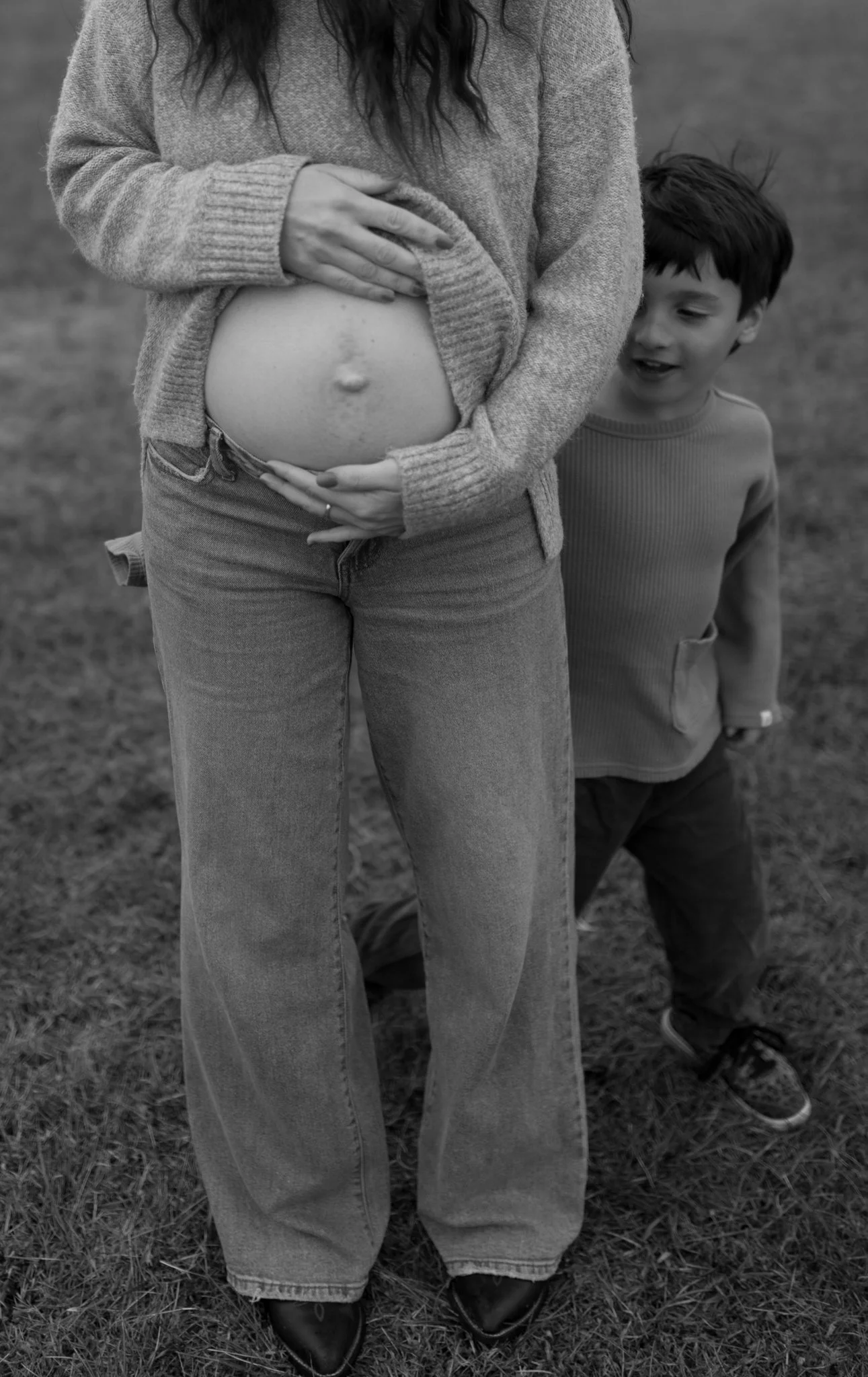 A pregnant woman holding her belly outdoors, with a young boy standing behind her and smiling.