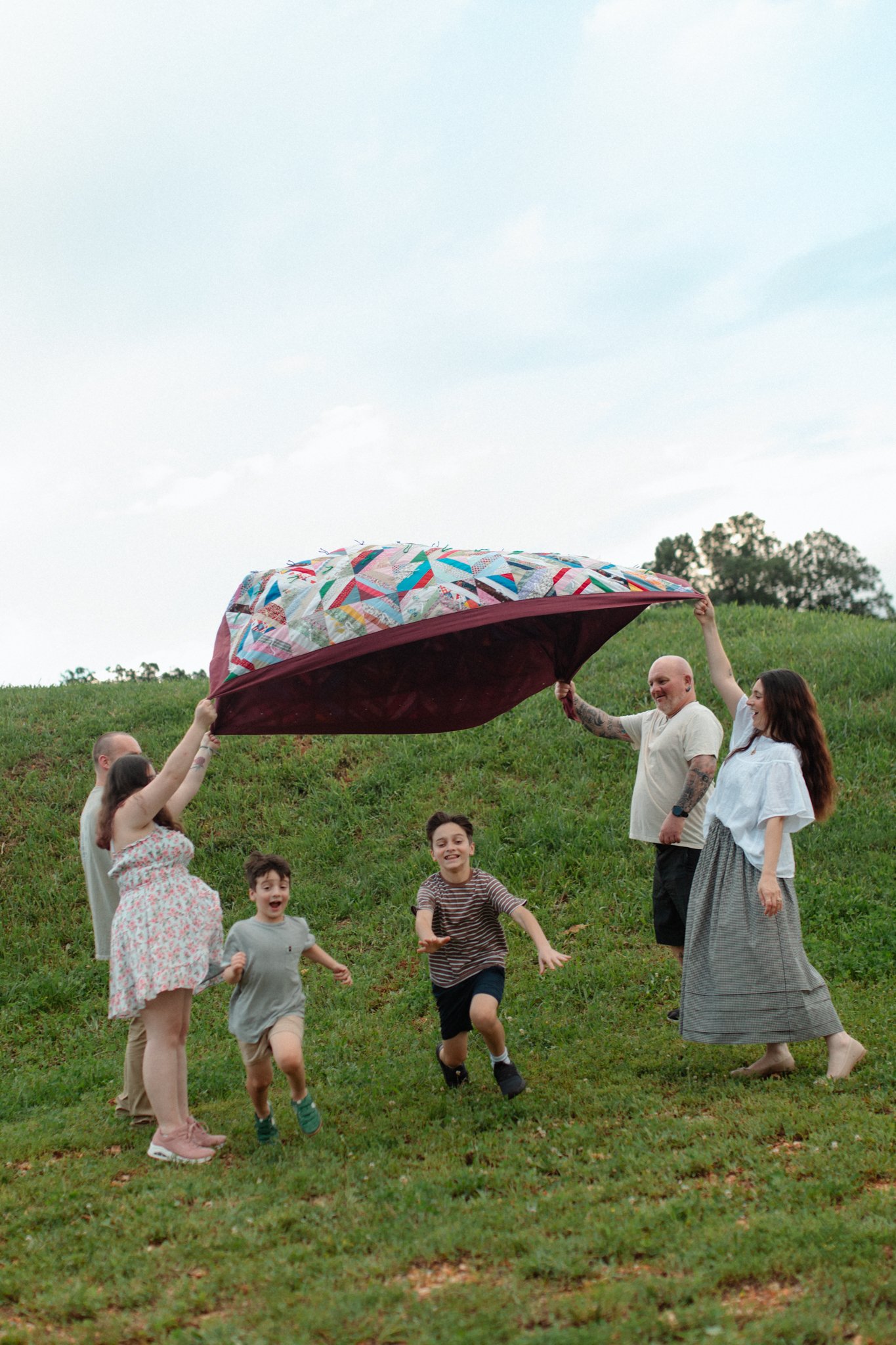 A family playing outdoors on a grassy hill with a large, colorful quilt.