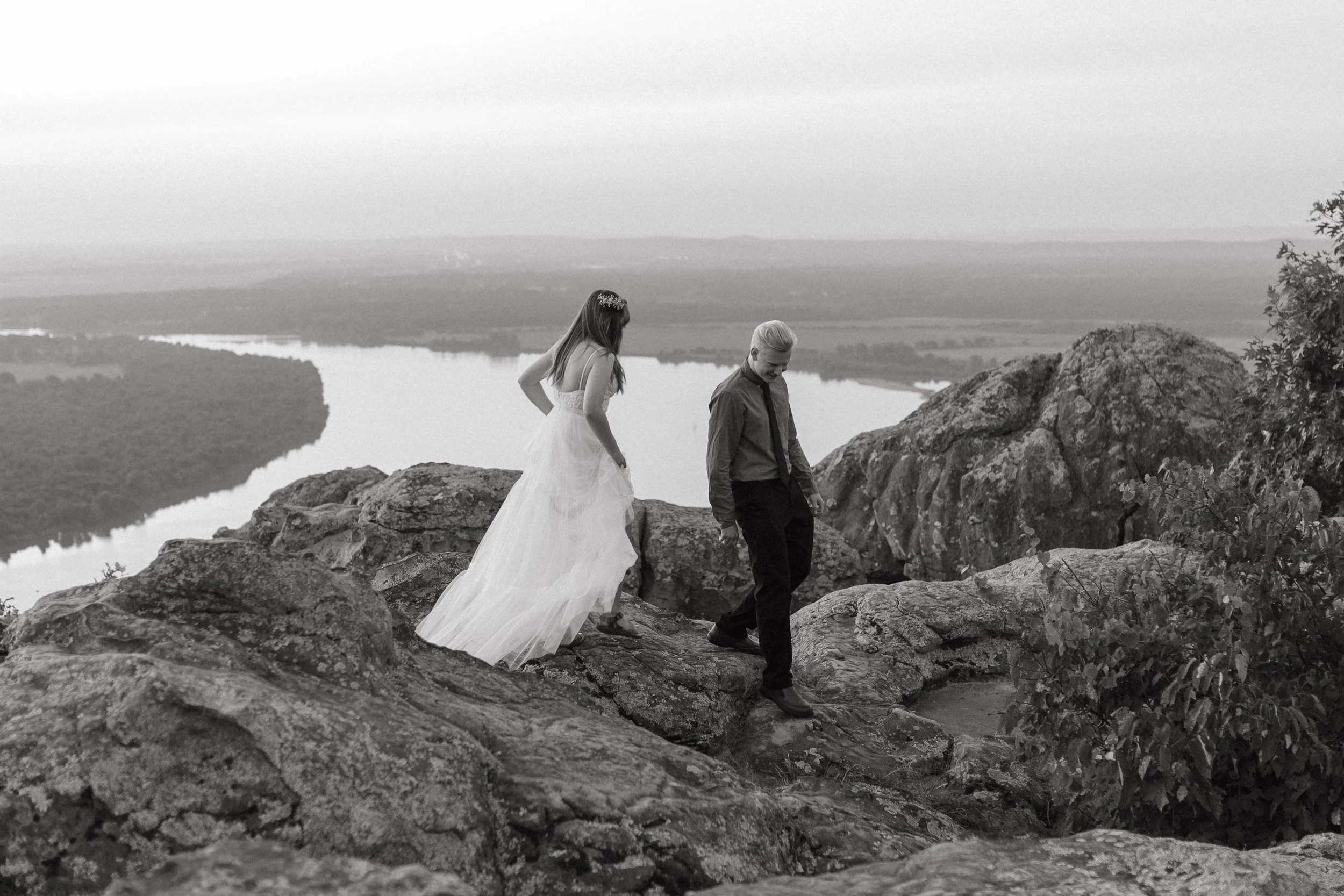 A woman in a long white dress and a man in a dark shirt and pants standing on rocks on a hilltop with a lake in the background, captured in black and white.