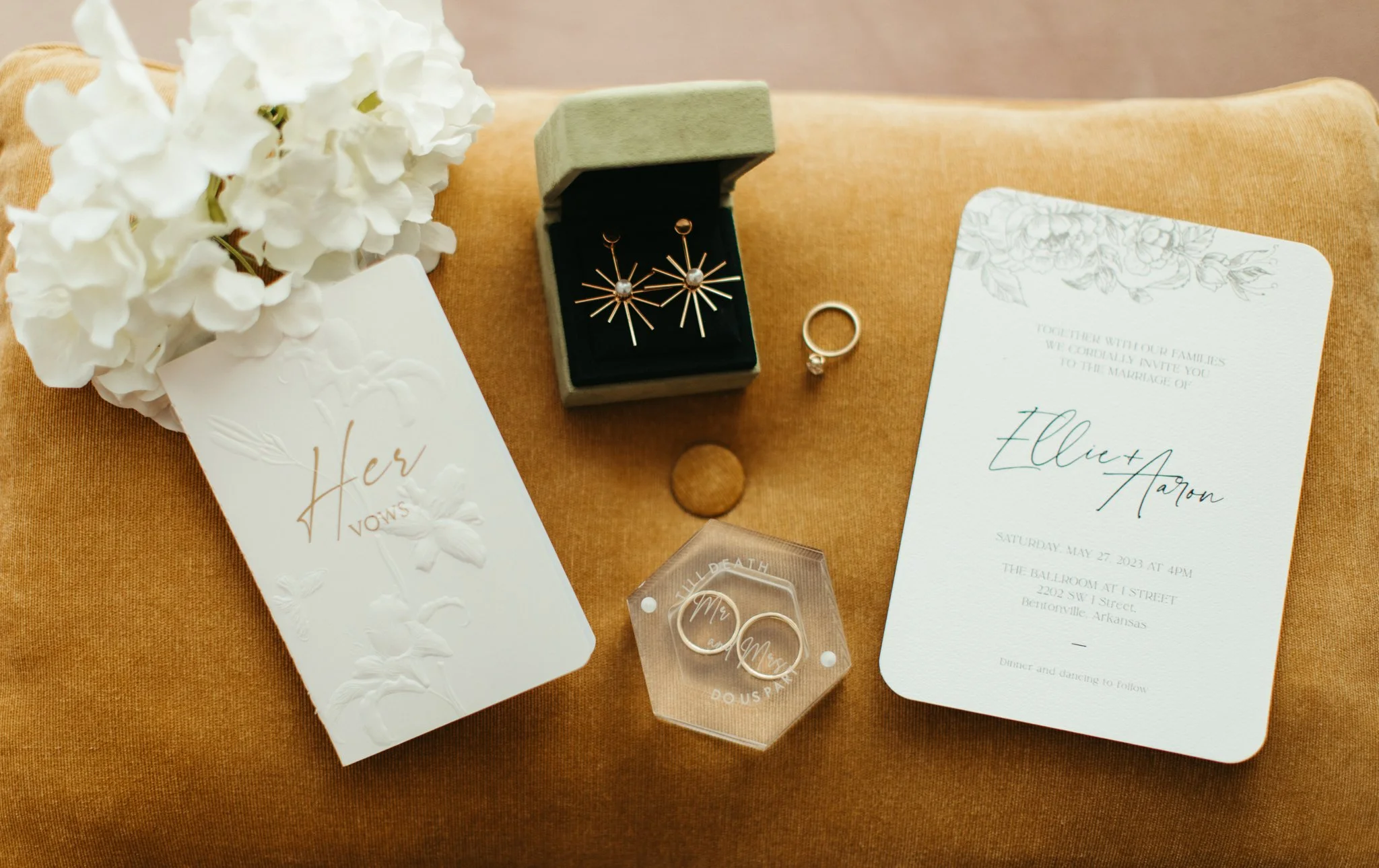 Wedding invitation, vows card, ring box, wedding rings, wedding rings in a clear case, earrings in a black box, flower arrangement, and a small round object on an orange cushion.