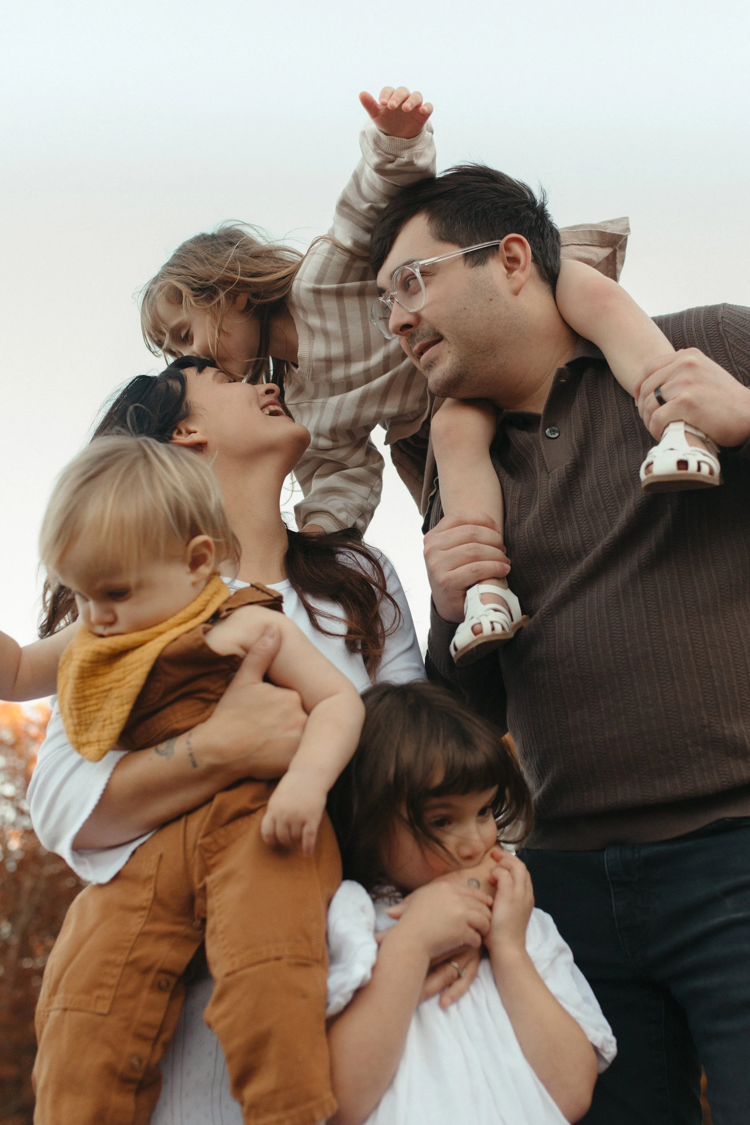 A family of six, including three adults and three children, are outdoors, sharing a playful moment. The adults are lifting children, and everyone is smiling and looking at each other affectionately.