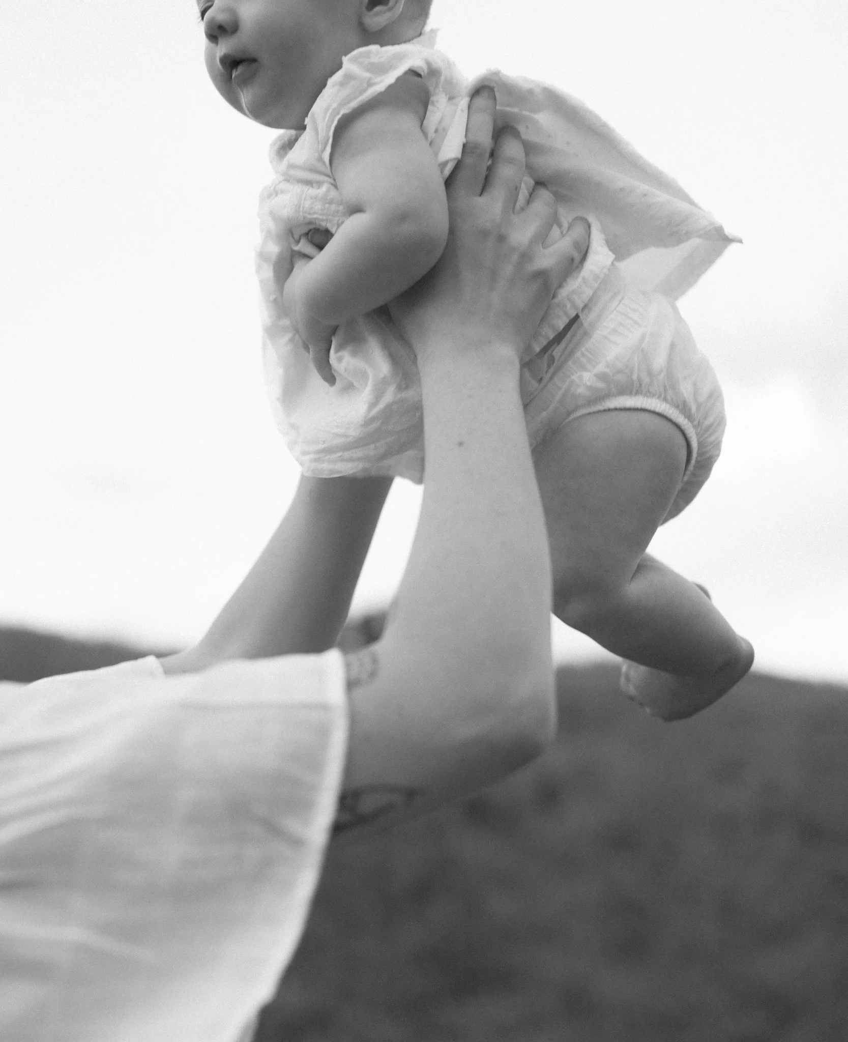 Black and white photo of a person lying on their back, holding a young child in the air. The child is wearing a cloth diaper and appears to be happily floating. The background is blurred, likely outdoors.