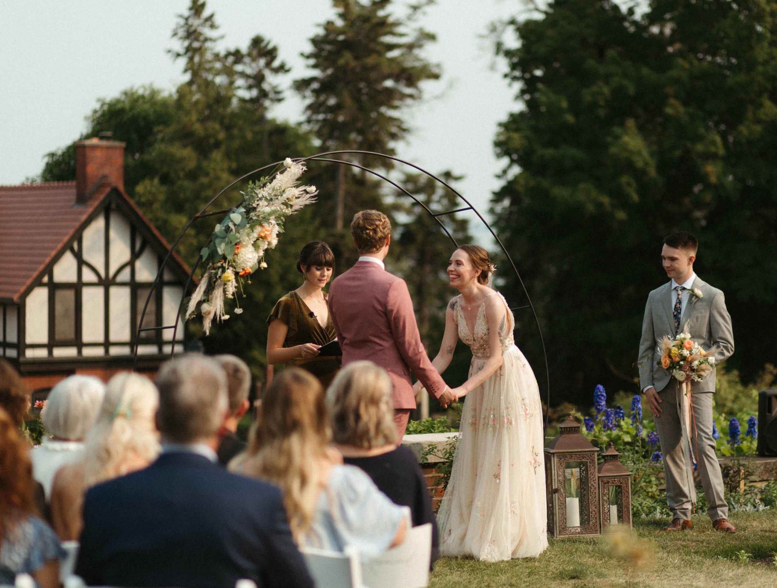 A couple exchanging vows during an outdoor wedding ceremony under a decorative arch, with guests seated and watching, and a man holding a bouquet standing nearby.