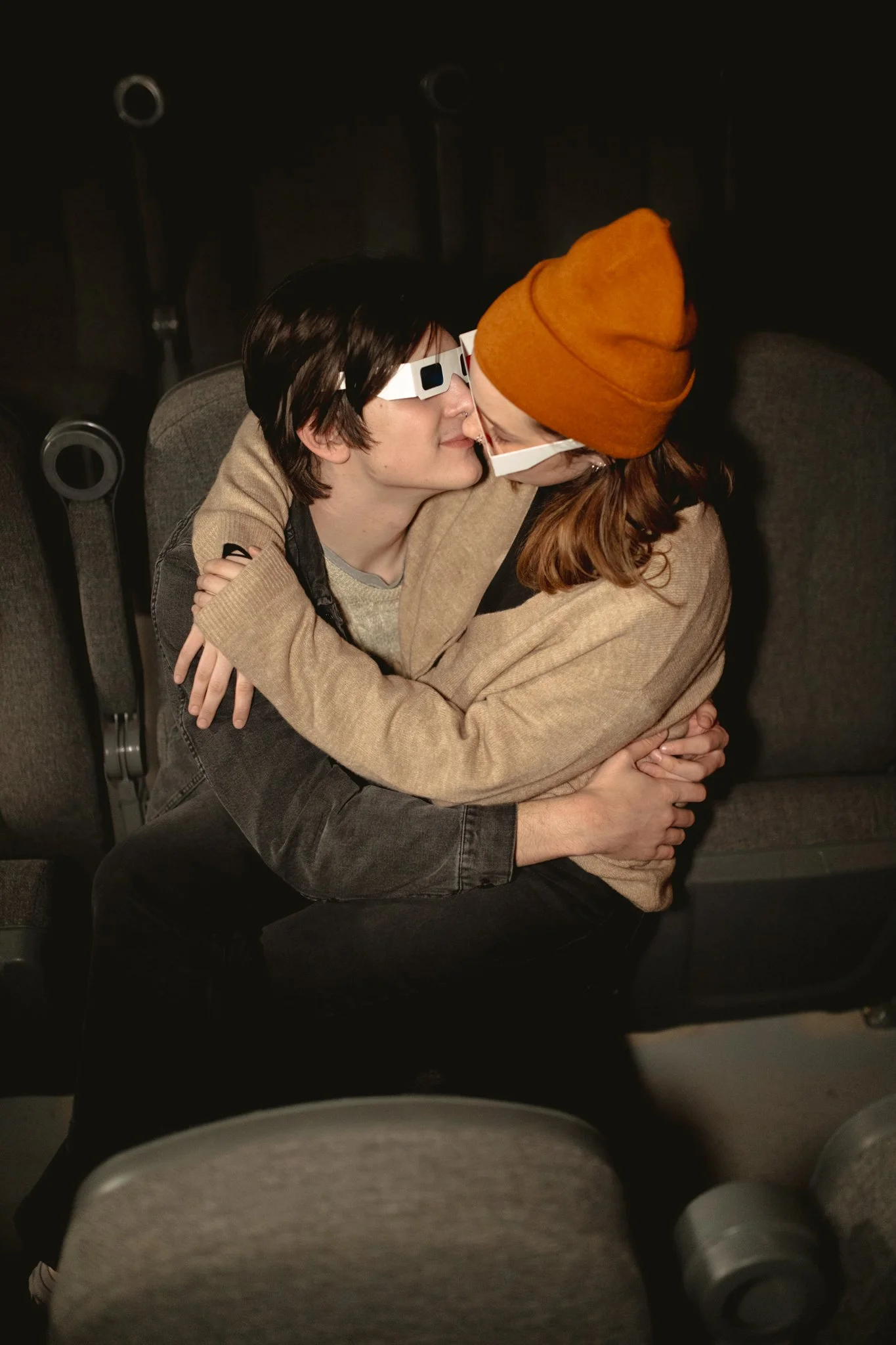 Two people are hugging and leaning in for a kiss in a dark theater, both wearing 3D glasses. One person has short dark hair and the other has long light brown hair, wearing an orange beanie and a face mask.