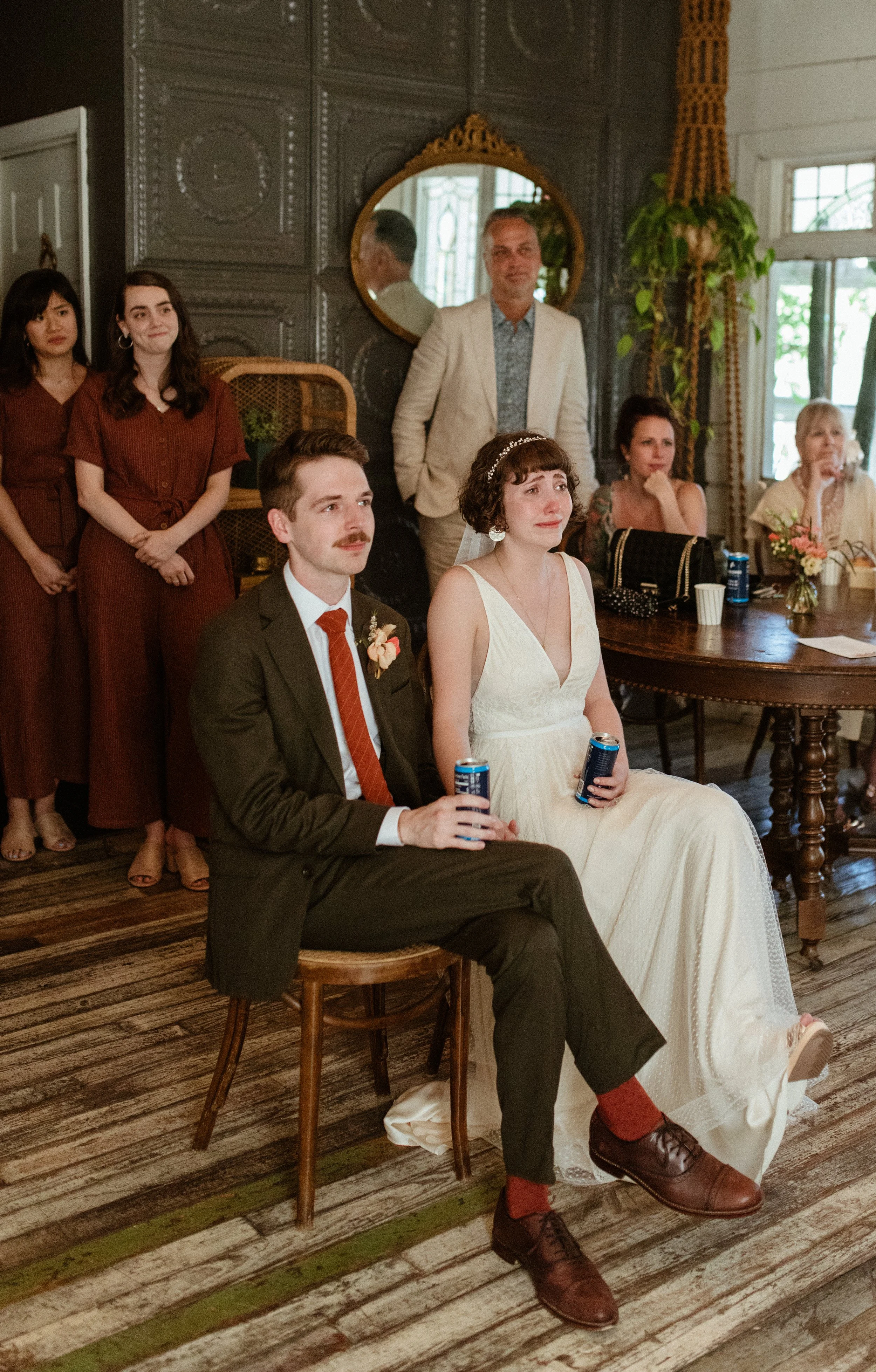 A bride and groom sit on chairs during a wedding ceremony, with guests standing and seated behind them, inside a decorated room with dark walls, a mirror, and large windows.