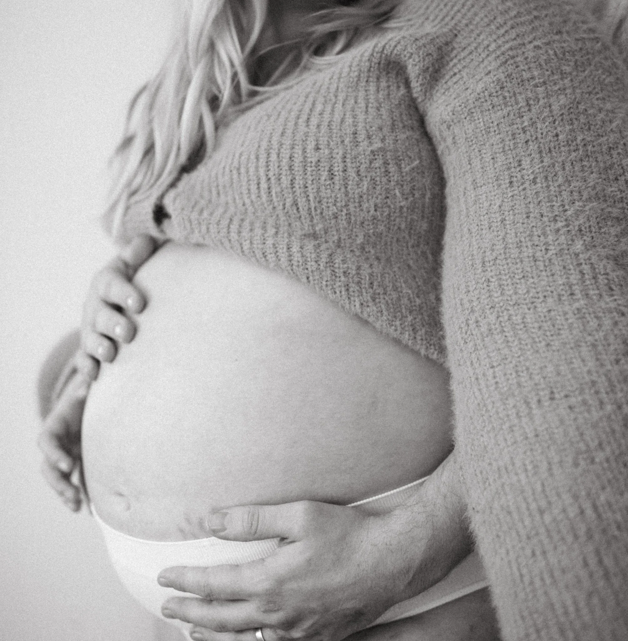 A pregnant woman cradling her belly with her hand, wearing a knitted sweater, in black and white.