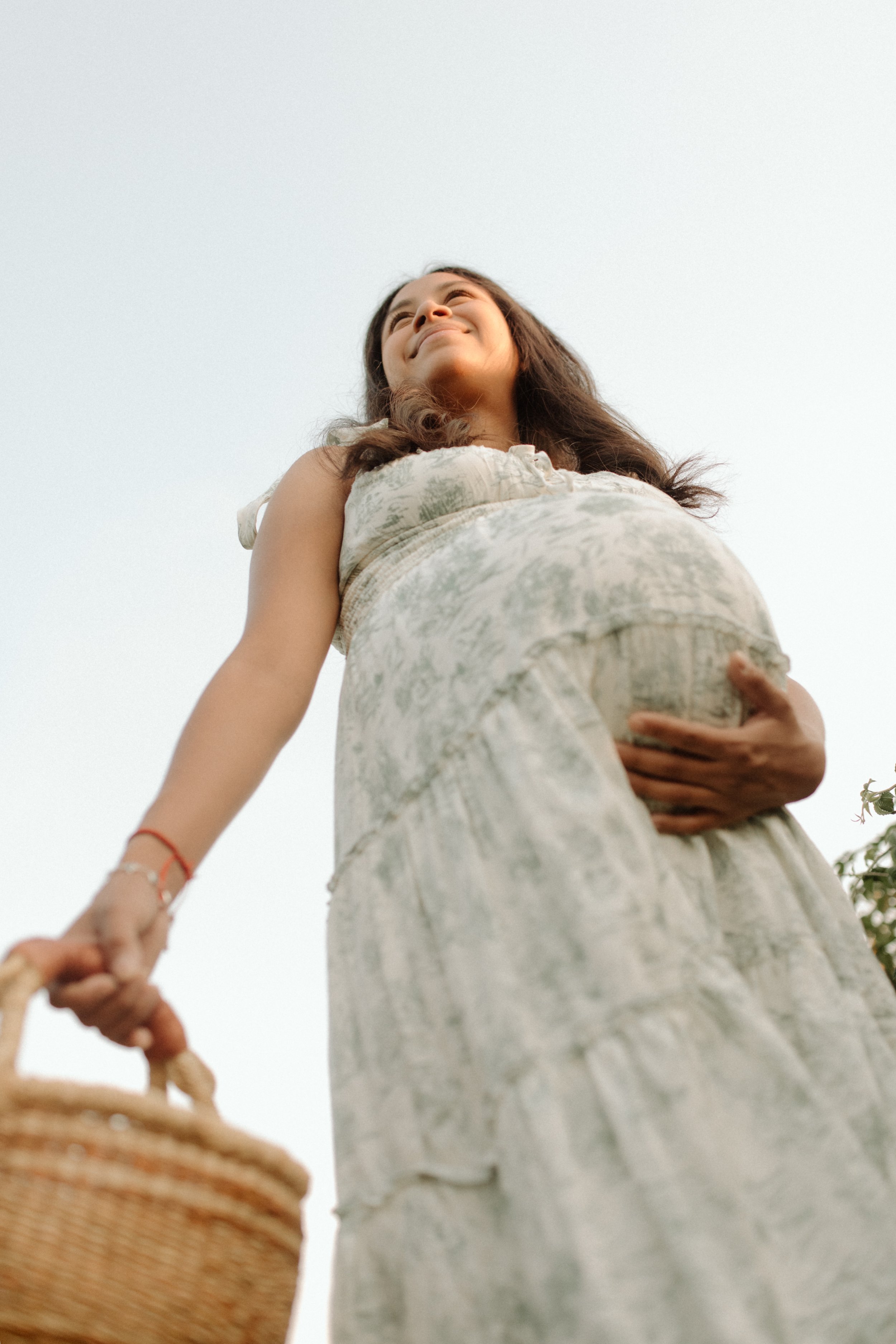 A pregnant woman in a light-colored dress smiling and holding her belly, with a basket in her right hand and outdoors with a clear sky.