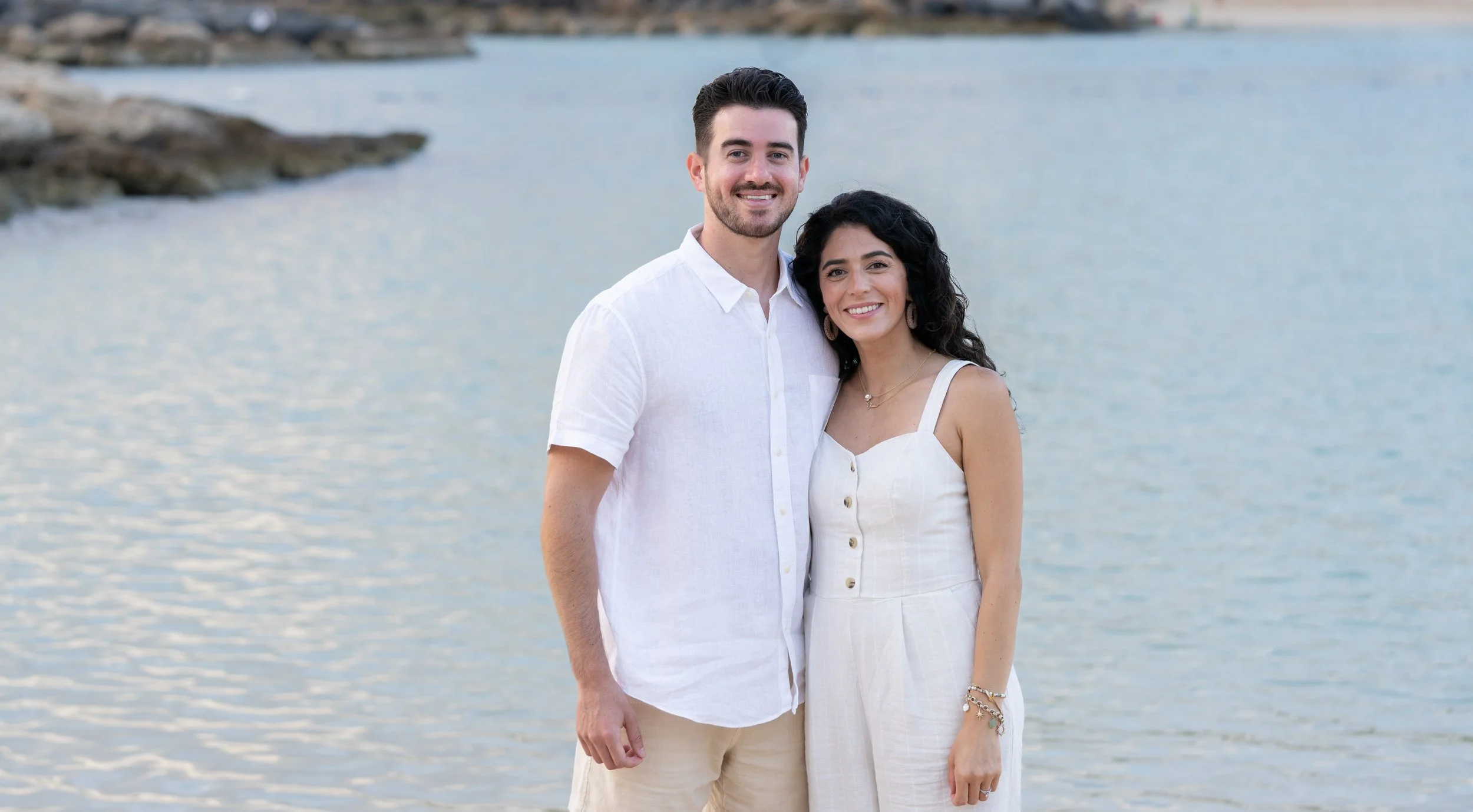 A smiling couple standing by the water, wearing light-colored summer clothing, with a rocky shoreline visible in the background.