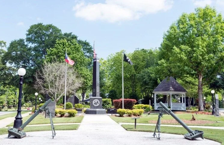 Memorial park with a central obelisk monument, American flags, a gazebo, and two large cannons on either side of the walkway.