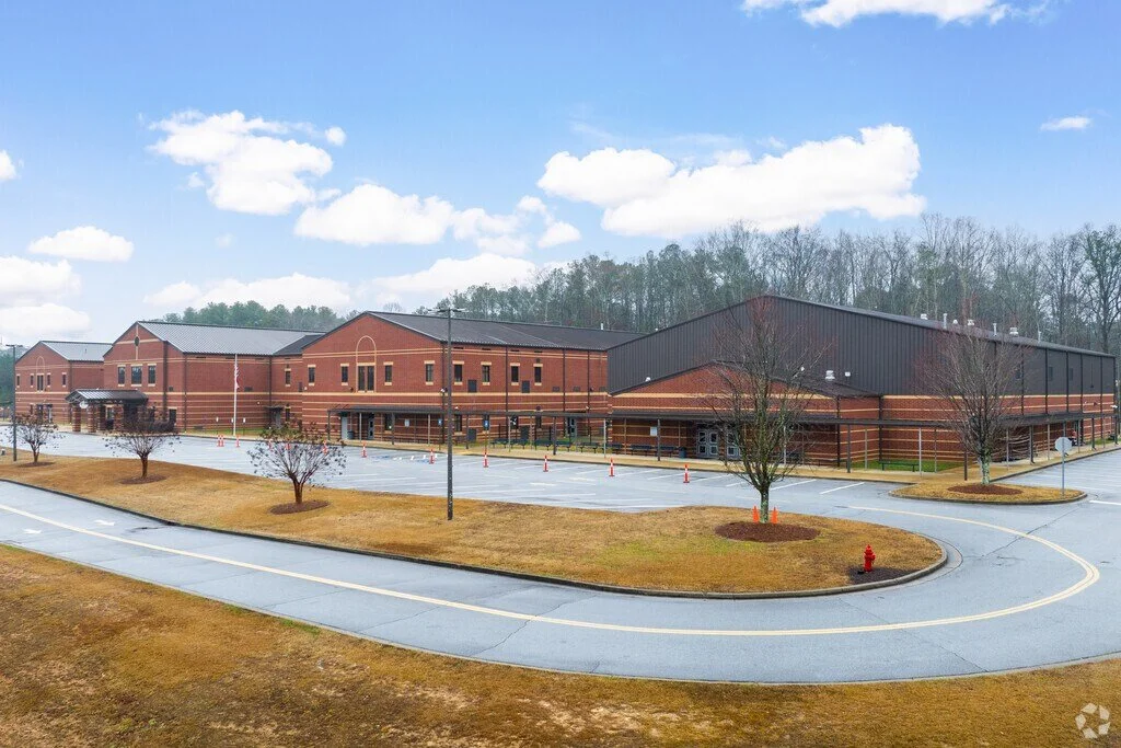 A large brick school building with a parking lot, surrounded by leafless trees, under a partly cloudy sky.