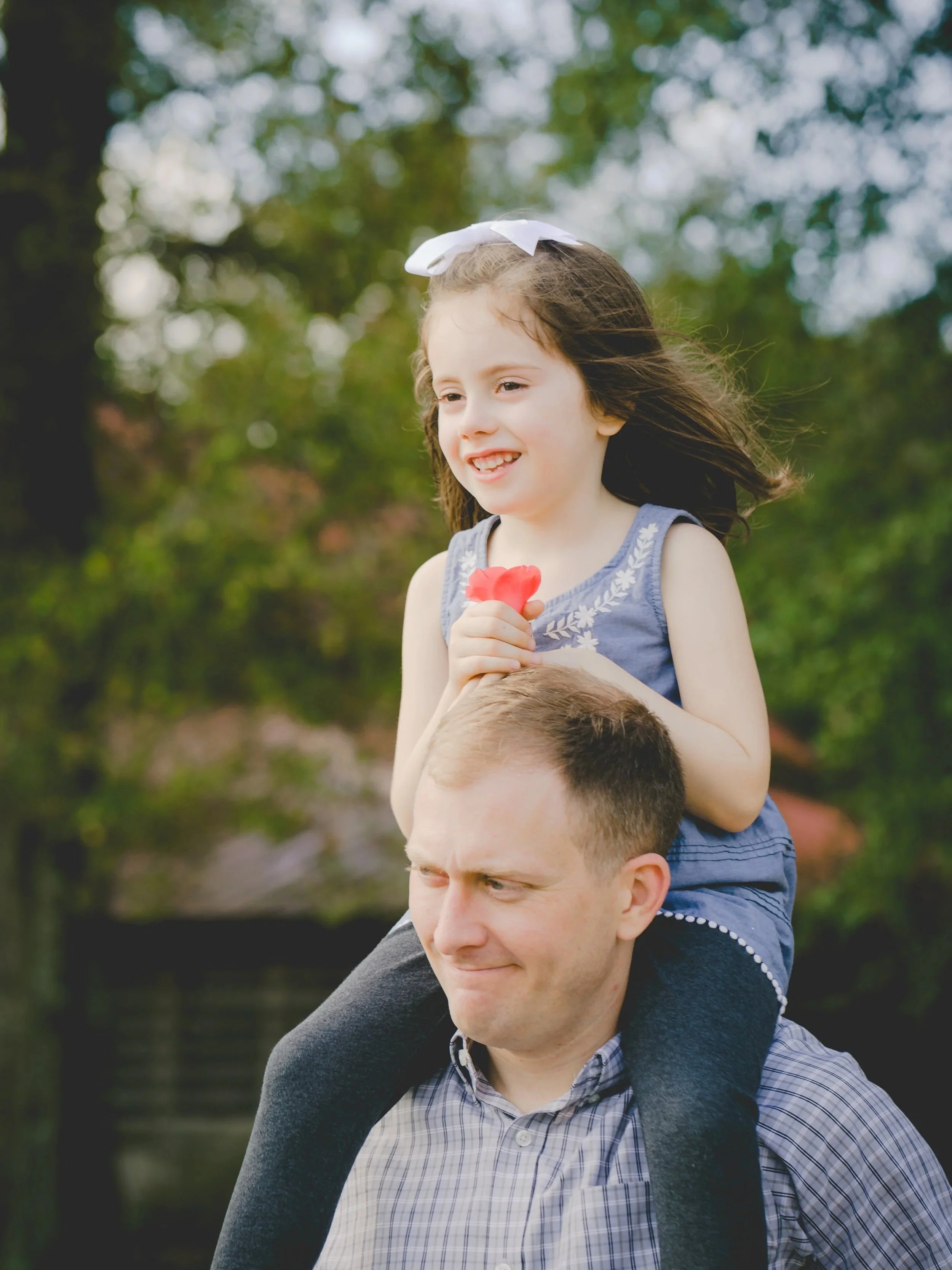 A young girl with a white bow in her hair riding on a man's shoulders outside in a park, holding a small red flower, with greenery and trees in the background.