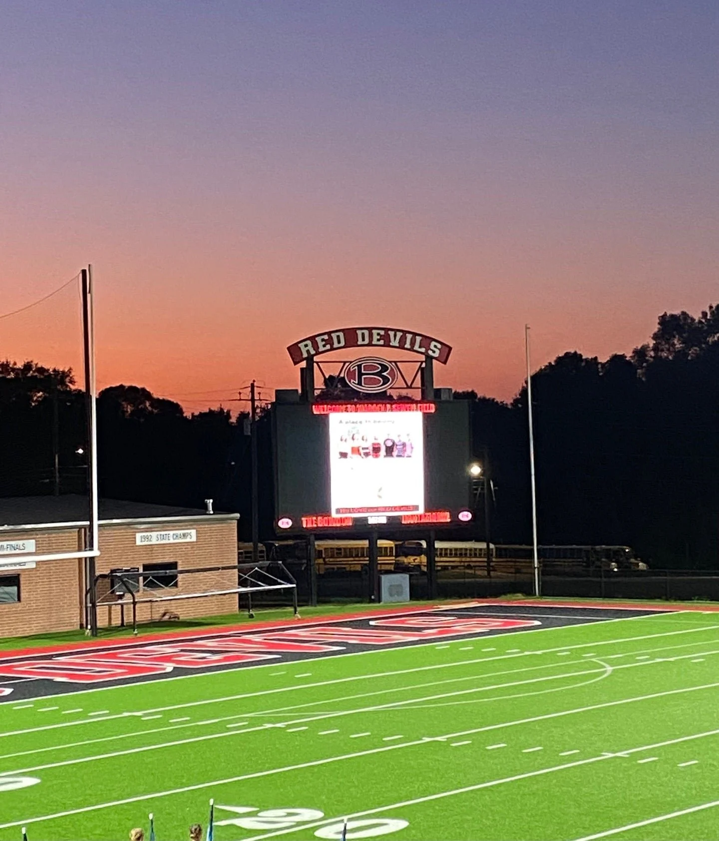 Football field at sunset with a digital scoreboard displaying the Red Devils logo and a message, surrounded by trees and sports facilities.