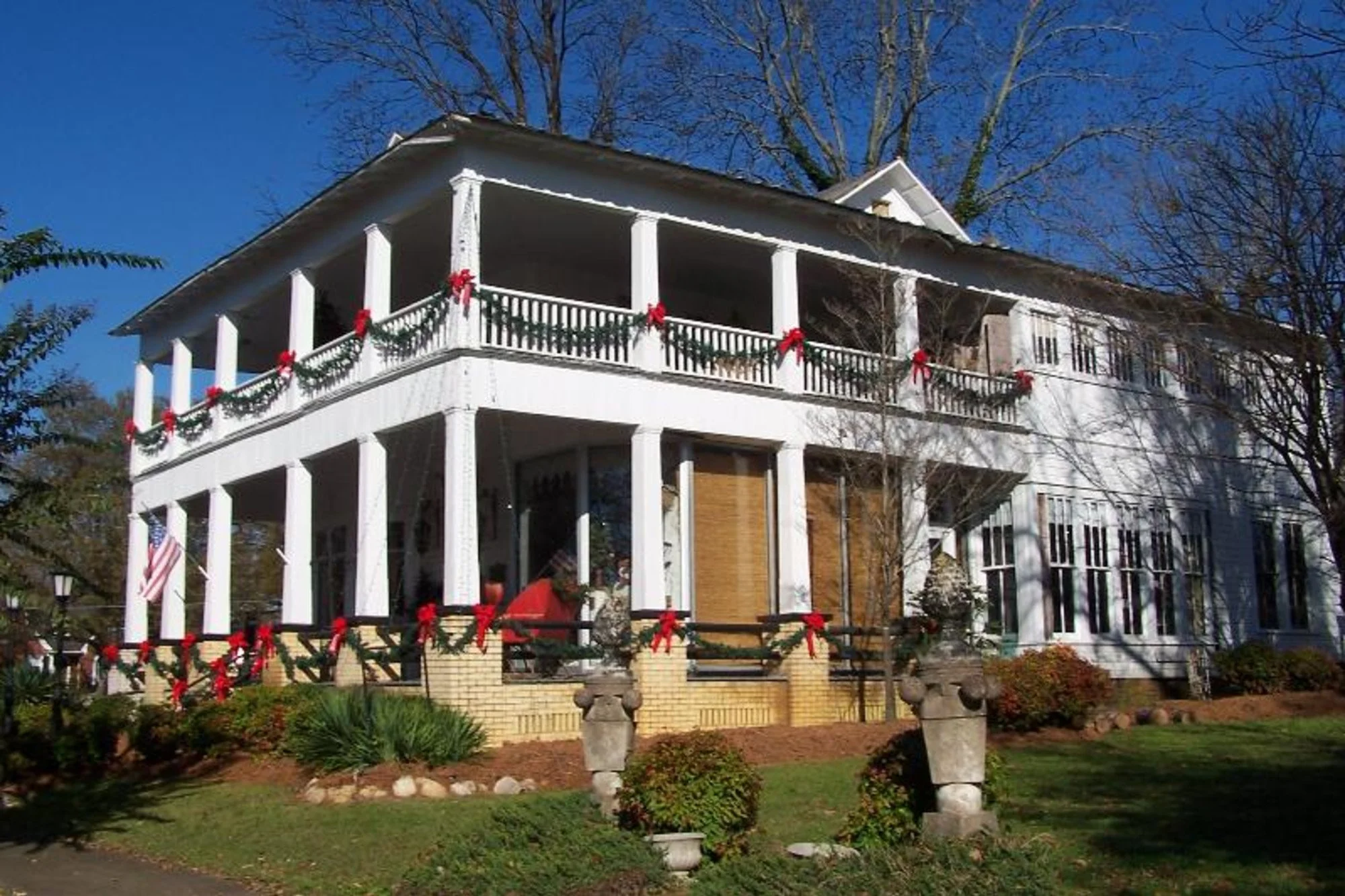 A white two-story house decorated with red bows and green garlands for Christmas, featuring a porch on both levels, with a light blue sky and trees in the background.
