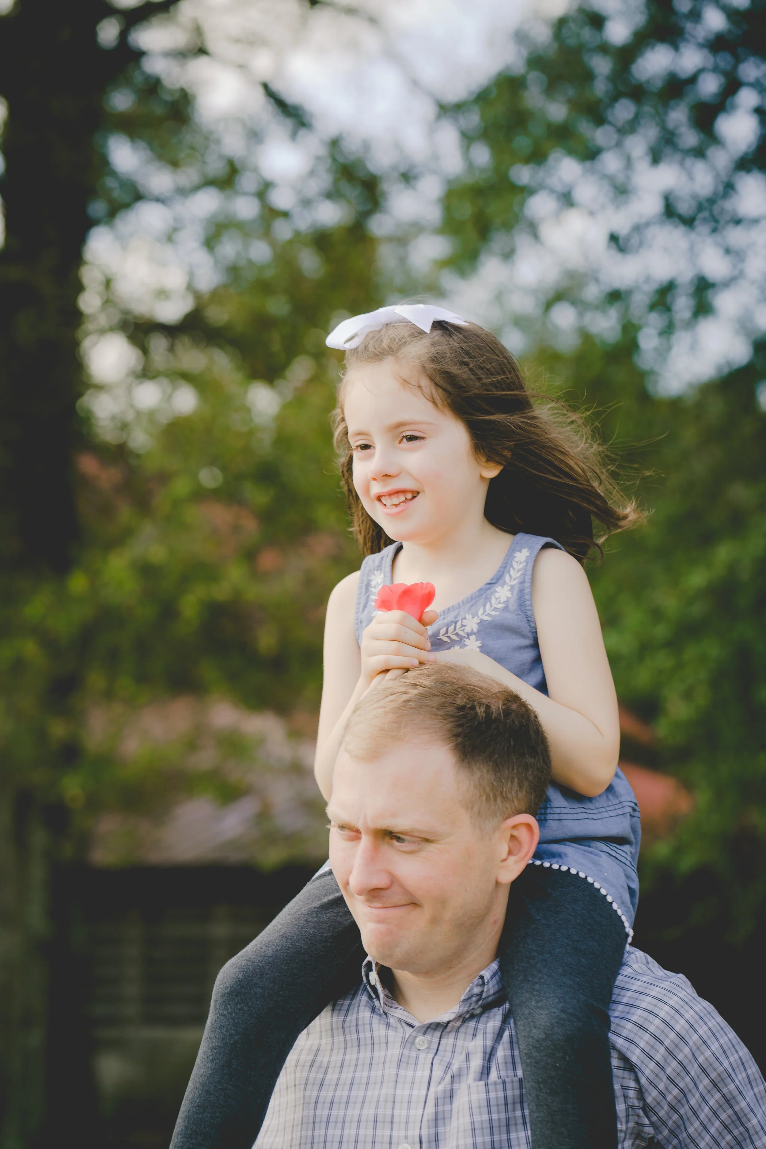 A young girl with brown hair wearing a blue dress and a white bow on her head is sitting on a man's shoulders outdoors. The girl is smiling and holding a pink flower, while the man, who has short hair and is wearing a plaid shirt, looks slightly downward with a content expression. The background features green trees and a slightly blurred natural setting.