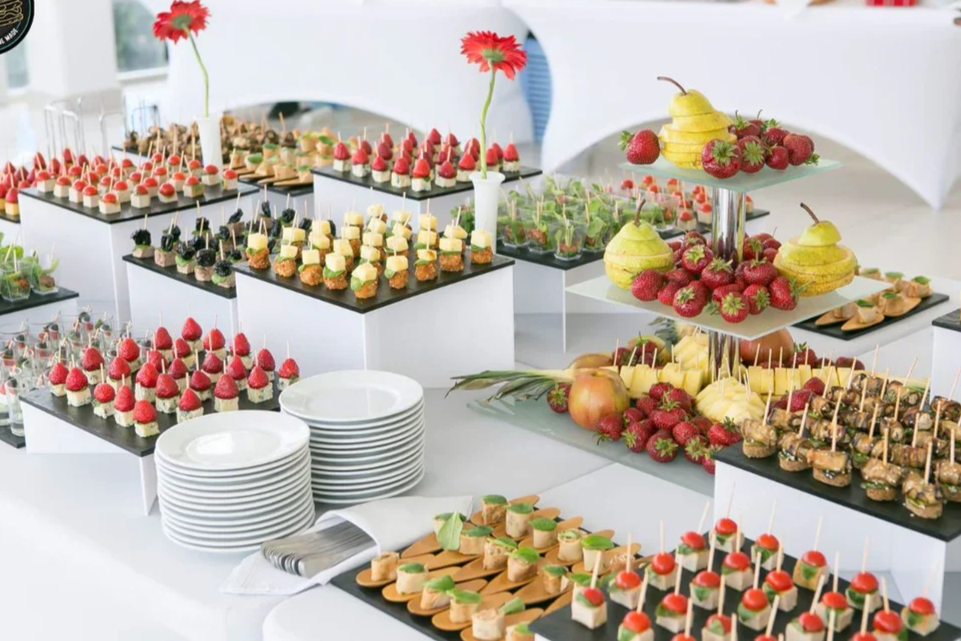 Display of assorted desserts and fruits on a table at a celebration, including small appetizer bites, strawberry and raspberry skewers, pears and strawberries, and stacks of plates.