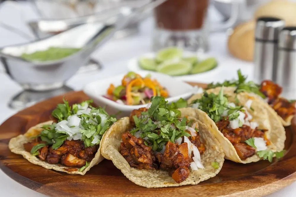 Three tacos filled with seasoned meat, topped with chopped onions and cilantro, served on a wooden platter with side dishes and condiments in the background.