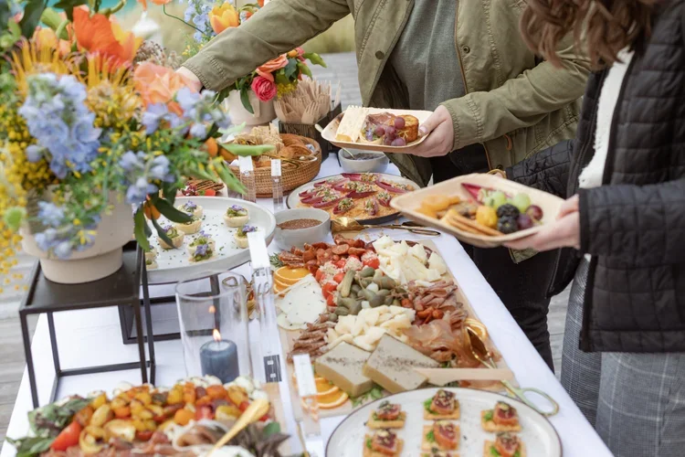 People serving themselves food at a buffet table with cheese, meats, fruits, and desserts, decorated with a colorful flower arrangement.