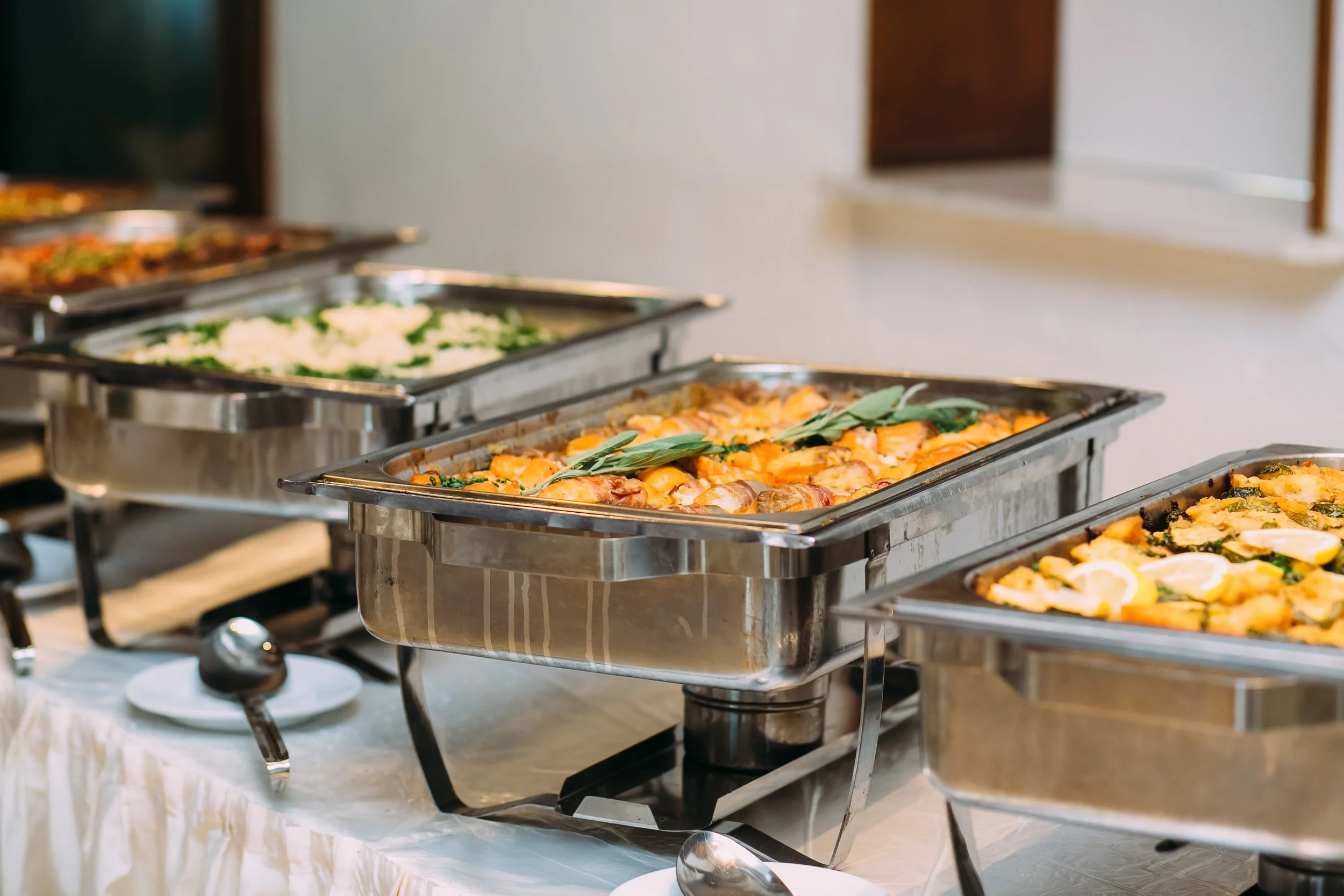 Buffet table with stainless steel chafing dishes containing various hot dishes, garnished with greens, set in a banquet or event setting.