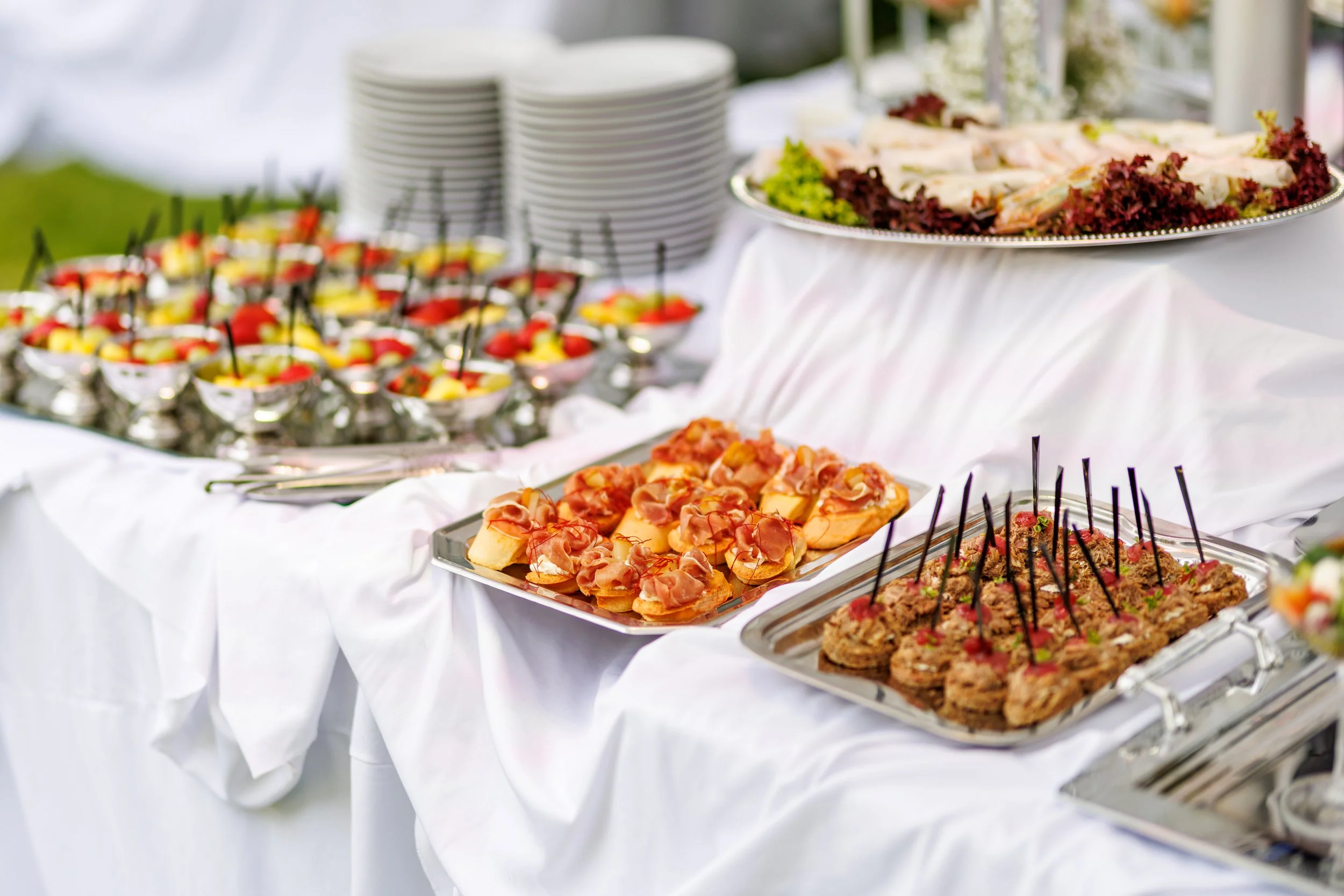 Buffet table with assorted appetizers including small cups of fruit and vegetables, sliced meats, and cheese on platters, covered with a white tablecloth.