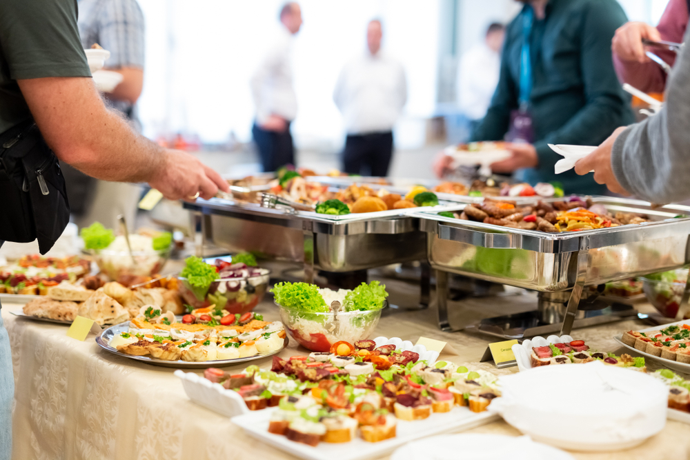Buffet table with various appetizers, salads, and finger foods in a bright room with people serving themselves.