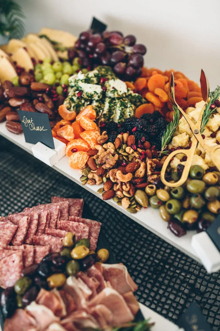 A display of assorted fruits, nuts, and cheeses on a white platter, including grapes, dried apricots, blackberries, candied nuts, and cheese slices