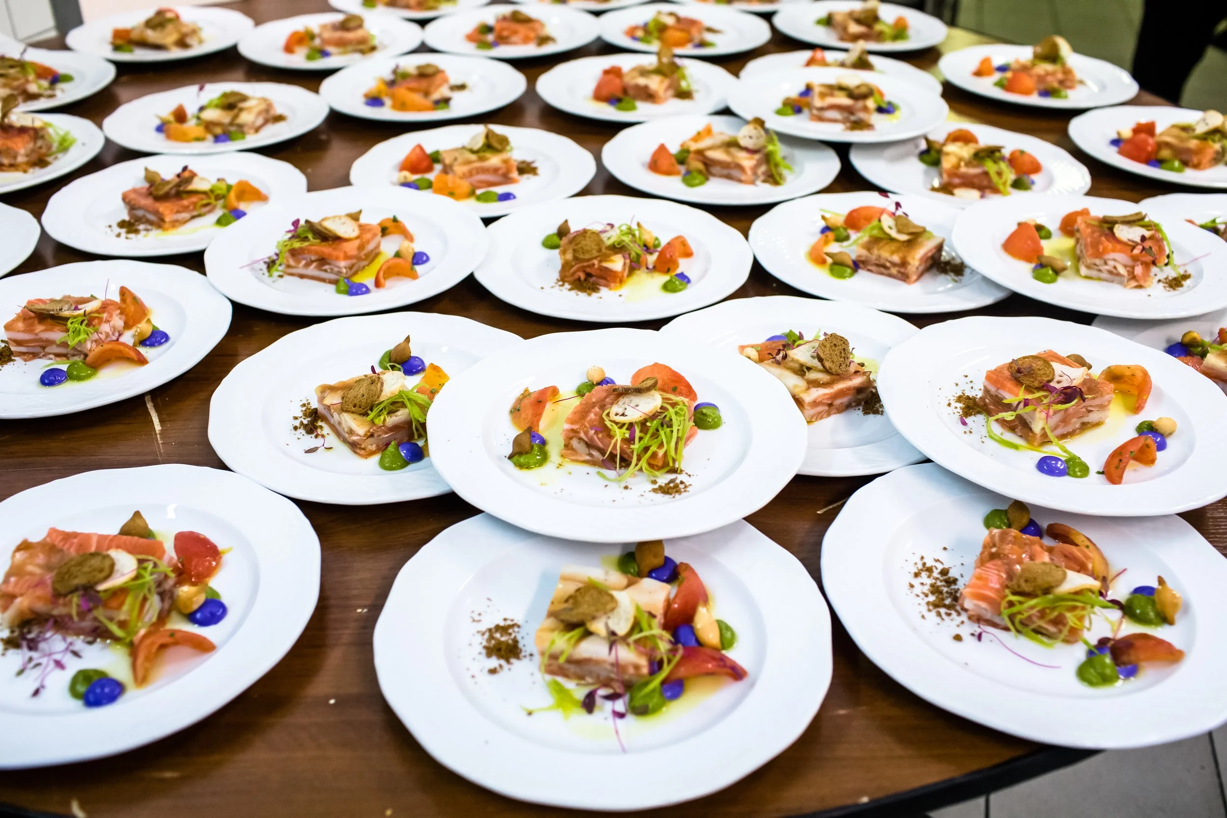 Multiple white plates with gourmet appetizer consisting of slices of salmon, garnished with green sprouts, small dollops of colorful sauces, and various herbs, arranged on a wooden table.