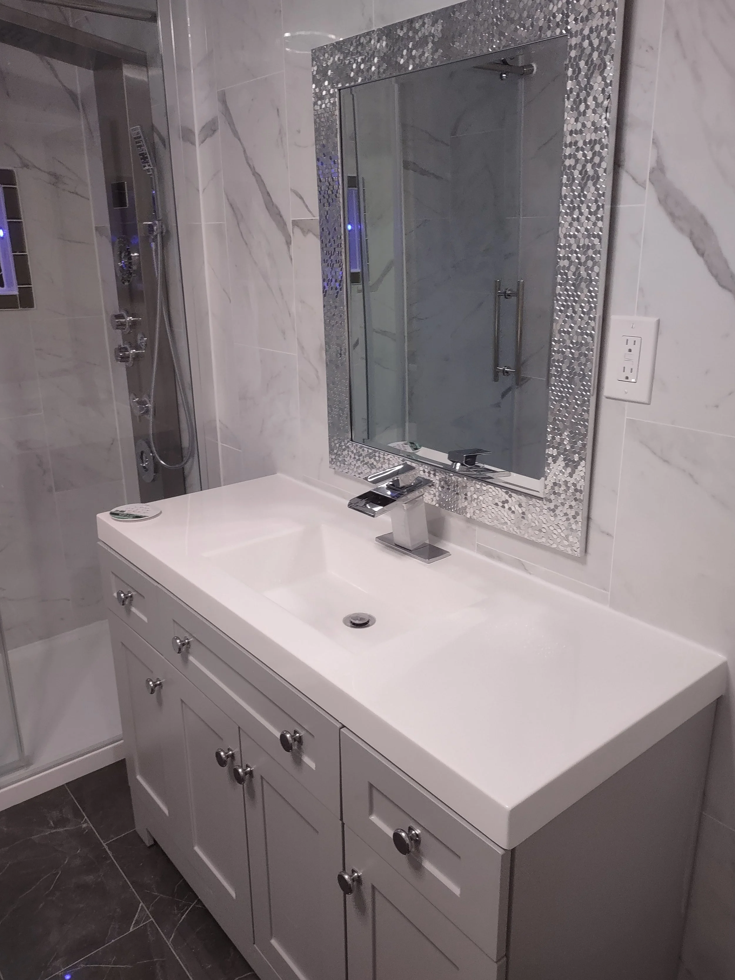 Modern bathroom with a white vanity, a rectangular sink, a sleek chrome faucet, and a large mirror with a decorative frame. In the background, there is a shower with glass doors and a wall-mounted shower panel with multiple jets.