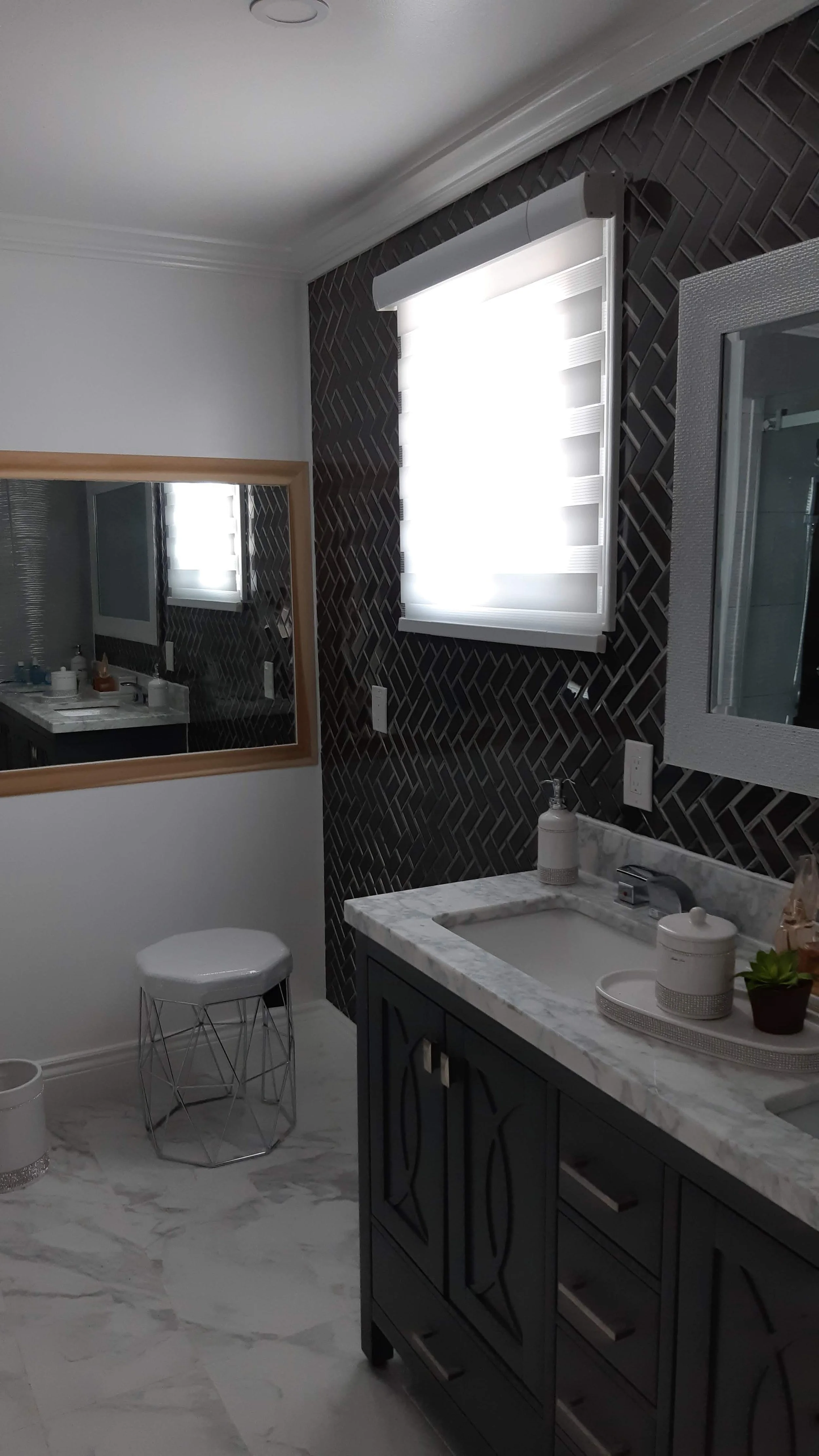 A modern bathroom with a dark tiled wall, a window with white blinds, a mirror, a marble countertop with a sink, soap dispenser, and decorative items, a dark wood vanity, a white stool, and a partial view of a large mirror reflecting the bathroom.