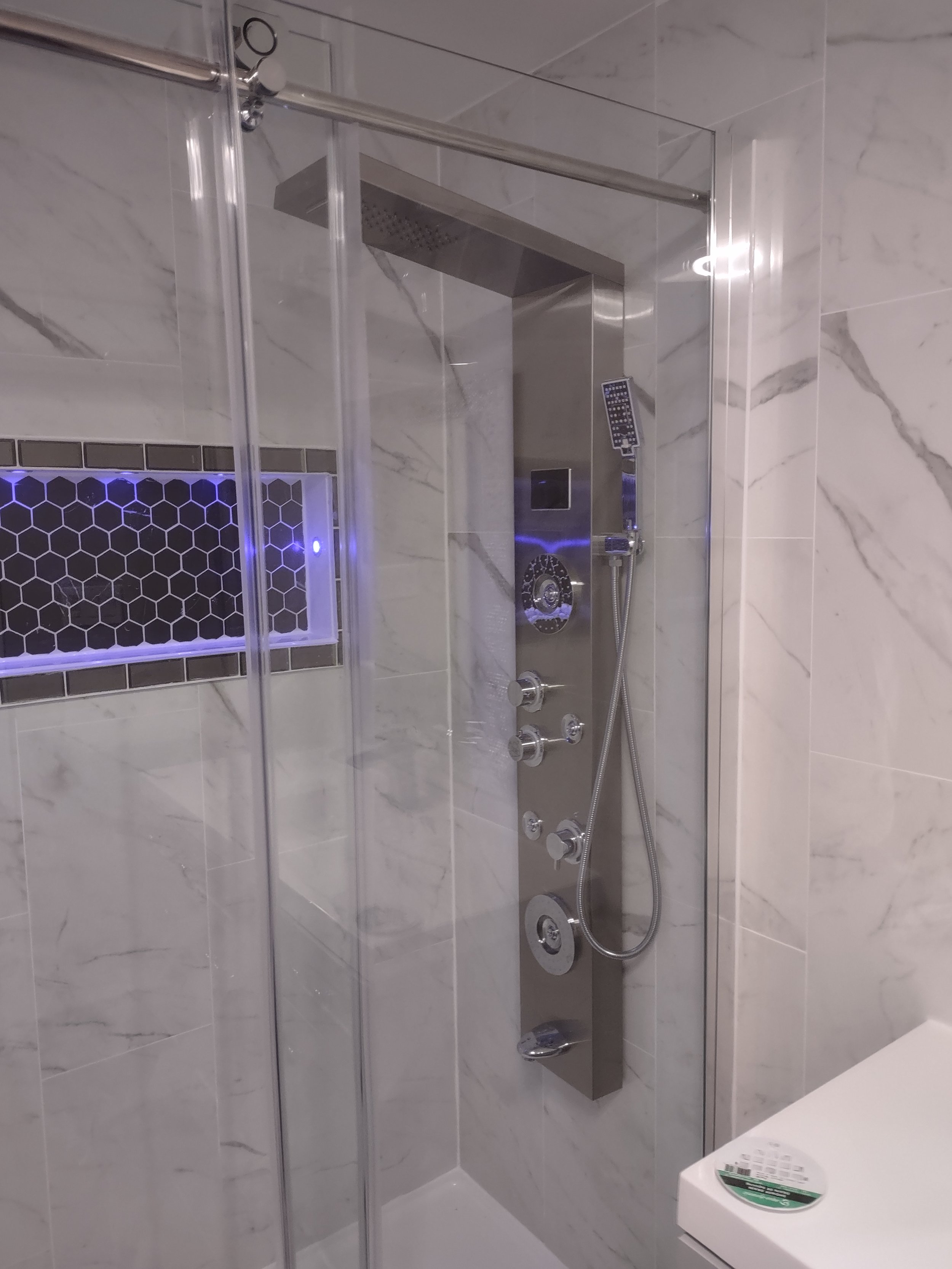 A modern shower with a glass enclosure, featuring a stainless steel panel with multiple showerheads, controls, and a handheld shower attachment in a marble-tiled bathroom.