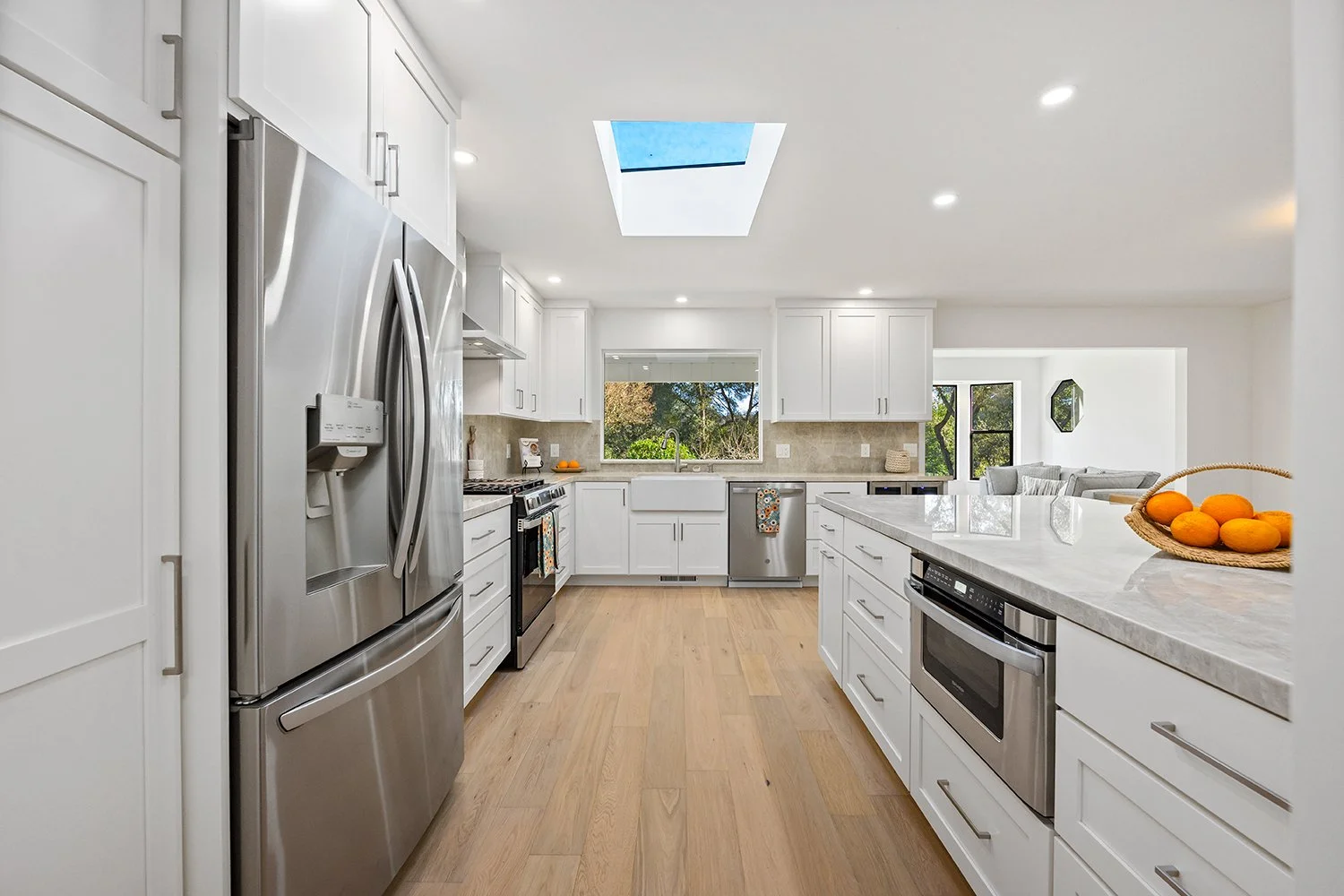 Modern kitchen with white cabinets, stainless steel appliances, wooden flooring, skylight, and large window showing greenery outside.
