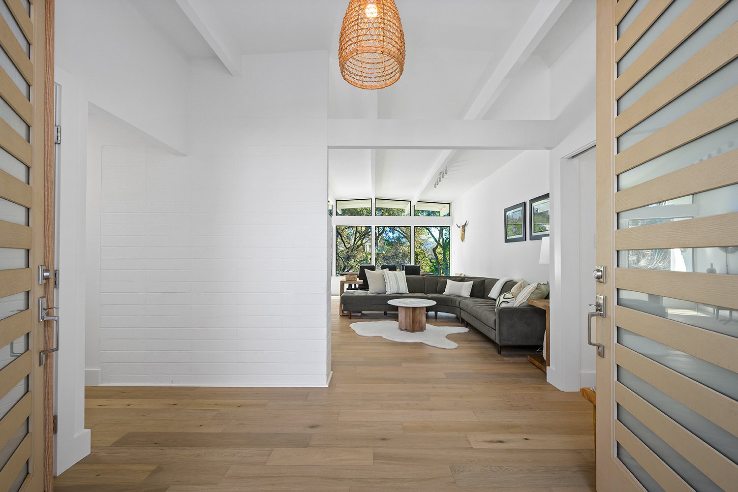View of a modern living room through an open doorway with wooden slatted doors. The room features a large L-shaped dark sofa with white and patterned pillows, a white cowhide rug, wooden floor, larger windows with trees outside, and framed pictures o
