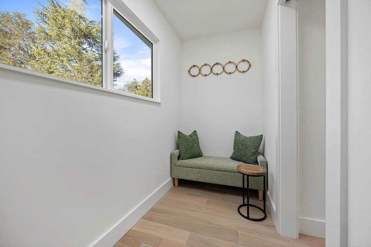A small cozy corner with a green upholstered bench with two green pillows, a small round side table with a wooden top and black metal frame, a white wall with a window showing trees outside, and a decorative wooden wall hanging.