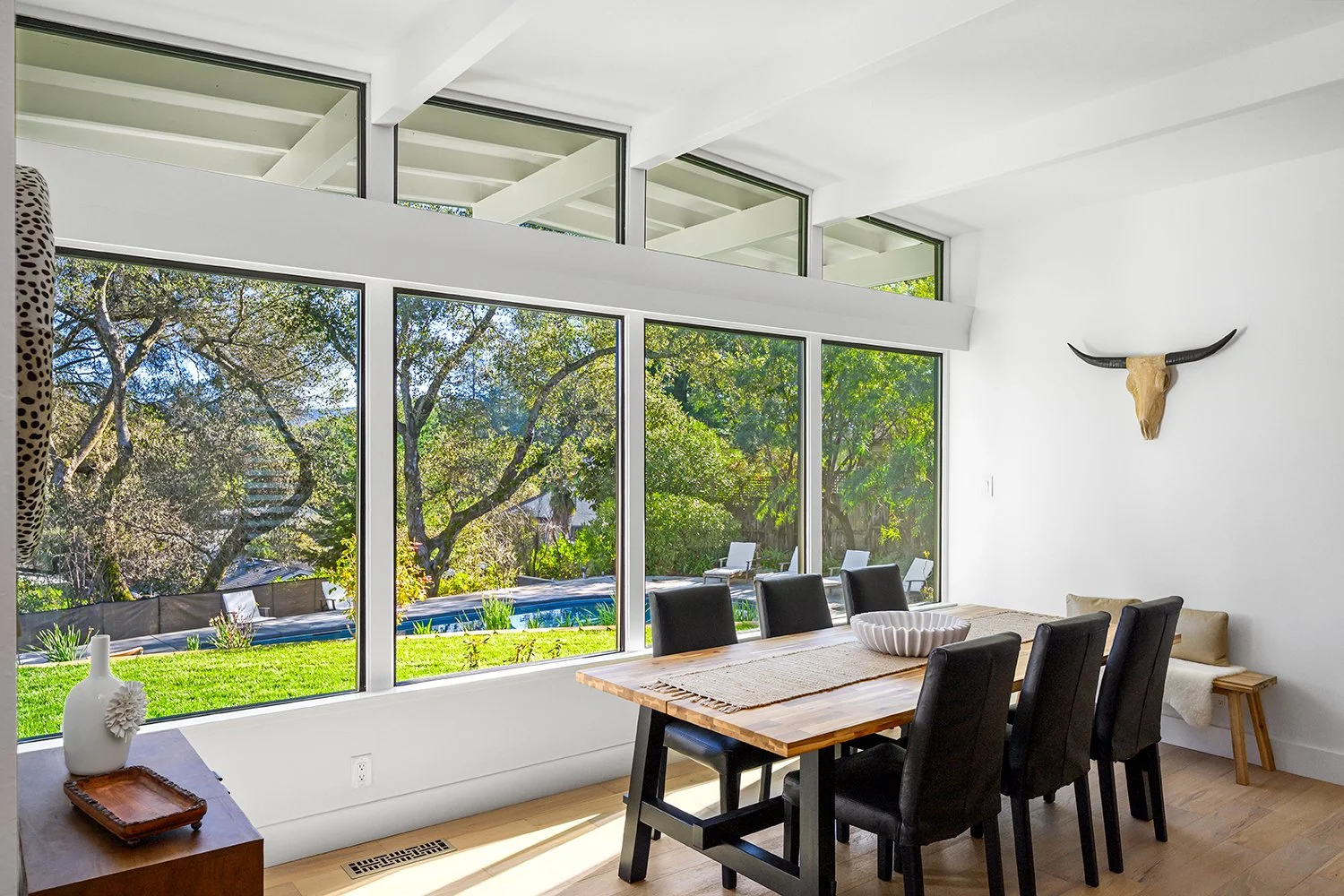 Dining room with large windows overlooking a backyard with trees and a pool, black chairs around a wooden table, decorative items on a side table, and a bull skull on the wall.