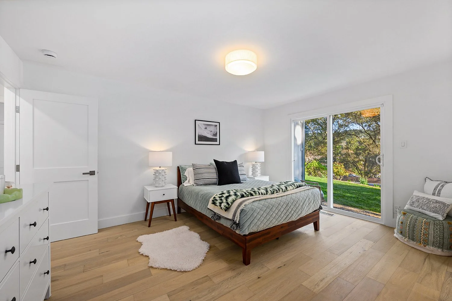 A bedroom with a wooden bed, white walls, and hardwood flooring. There are two white bedside tables with lamps, a small framed picture above the bed, and a sliding glass door leading outside to a green yard with trees.