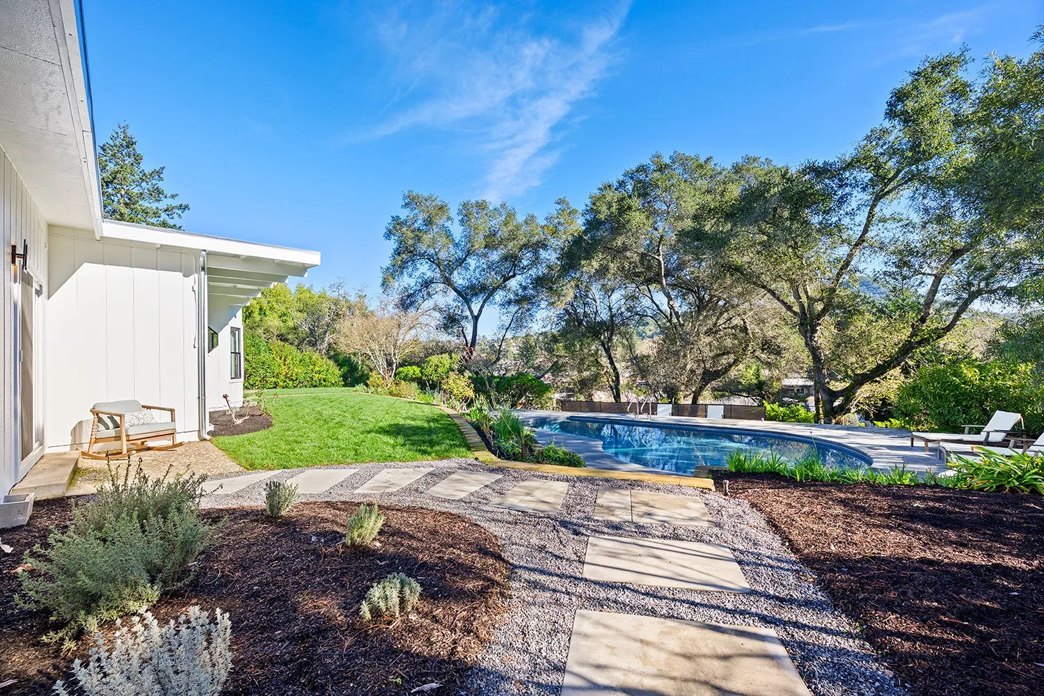 A backyard with a lawn, a swimming pool surrounded by trees, and a patio with chairs and a bench near a white house under a blue sky.