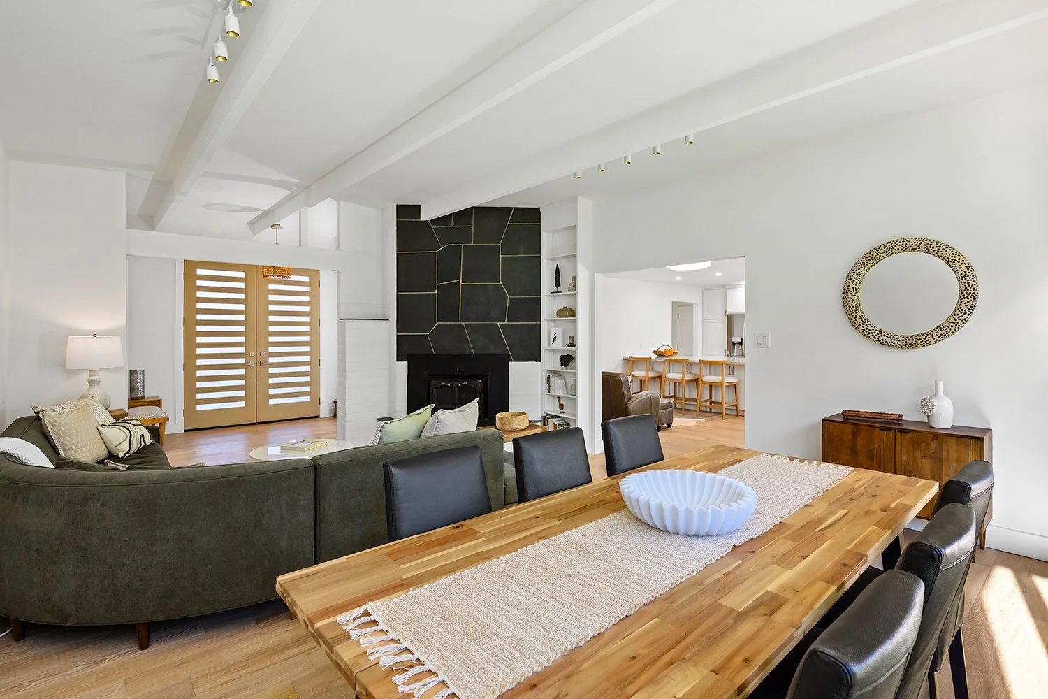 Spacious living and dining area with wooden flooring, black and green couch, wooden dining table with black chairs, fireplace with black tiled wall, white shelving, and decorative items.