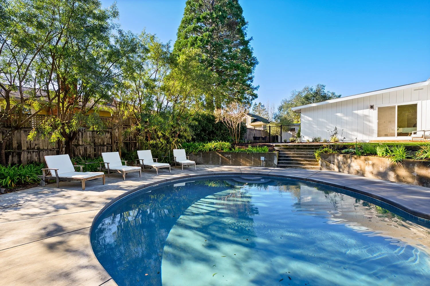 Backyard with an in-ground swimming pool, four lounge chairs, trees, plants, steps, and a white house with sliding glass door under a clear blue sky.