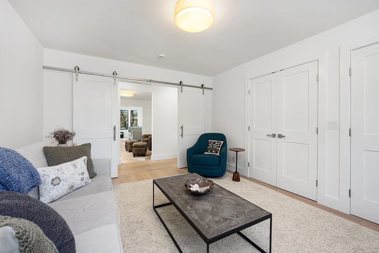 Living room with white walls, sliding barn doors, beige sofa with pillows, blue armchair with decorative pillow, coffee table, nearby side table, and view into another room with large window and armchair.