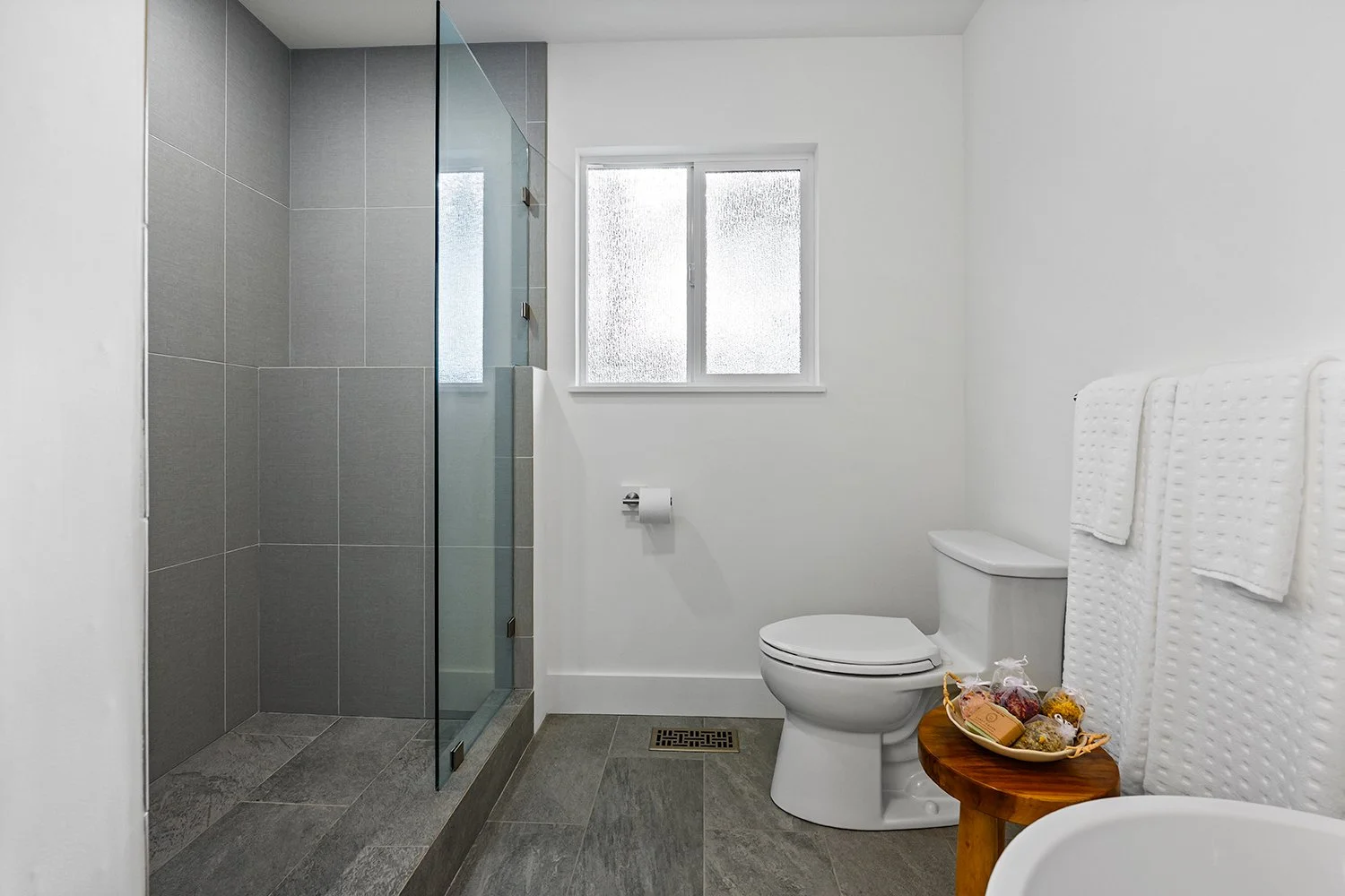 Bathroom with shower, toilet, and window, with white towels hanging on a rack and a small wooden table with a basket of soaps and bath products.