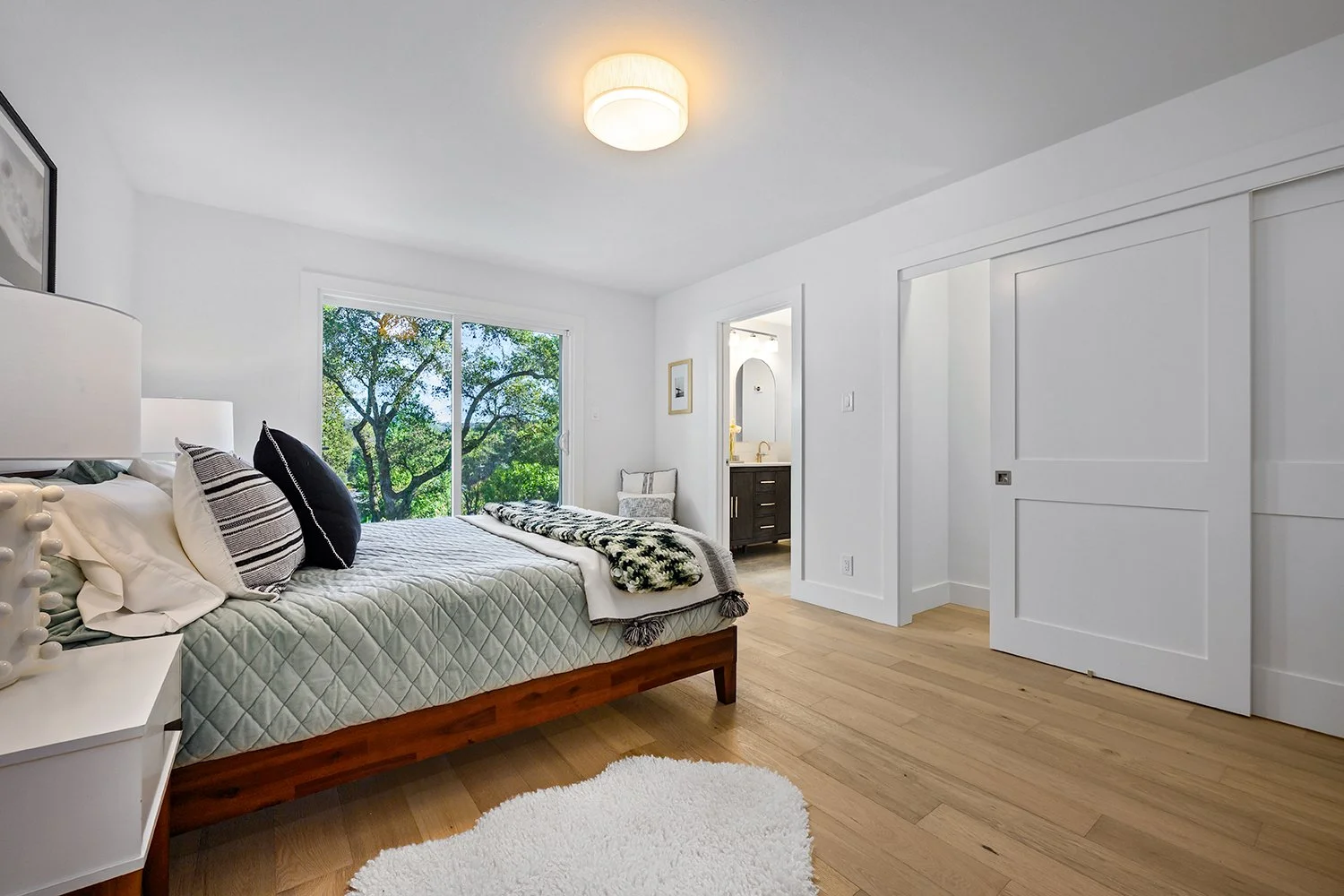 Bright bedroom with white walls, wooden bed frame, white quilt, and black and white pillows. Large window with trees outside, small white side table, and a white shag rug on wooden floor. Bathroom visible through doorway.