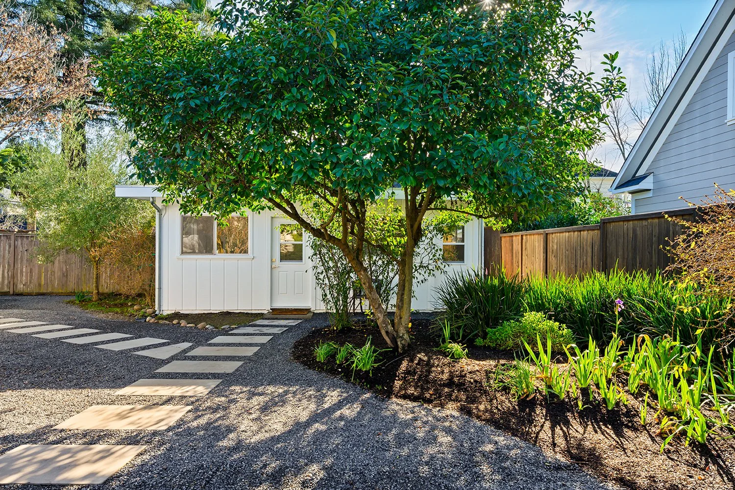 A small white house with a white door and windows, surrounded by greenery, including a large tree in front, on a gravel pathway with concrete stepping stones, in a backyard with a wooden fence and neighboring white house.