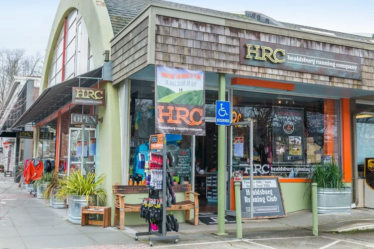 Exterior view of the HRC headsburg running company store with clothing displays outside, including jackets and socks, a bench, and a handicap parking sign in front.