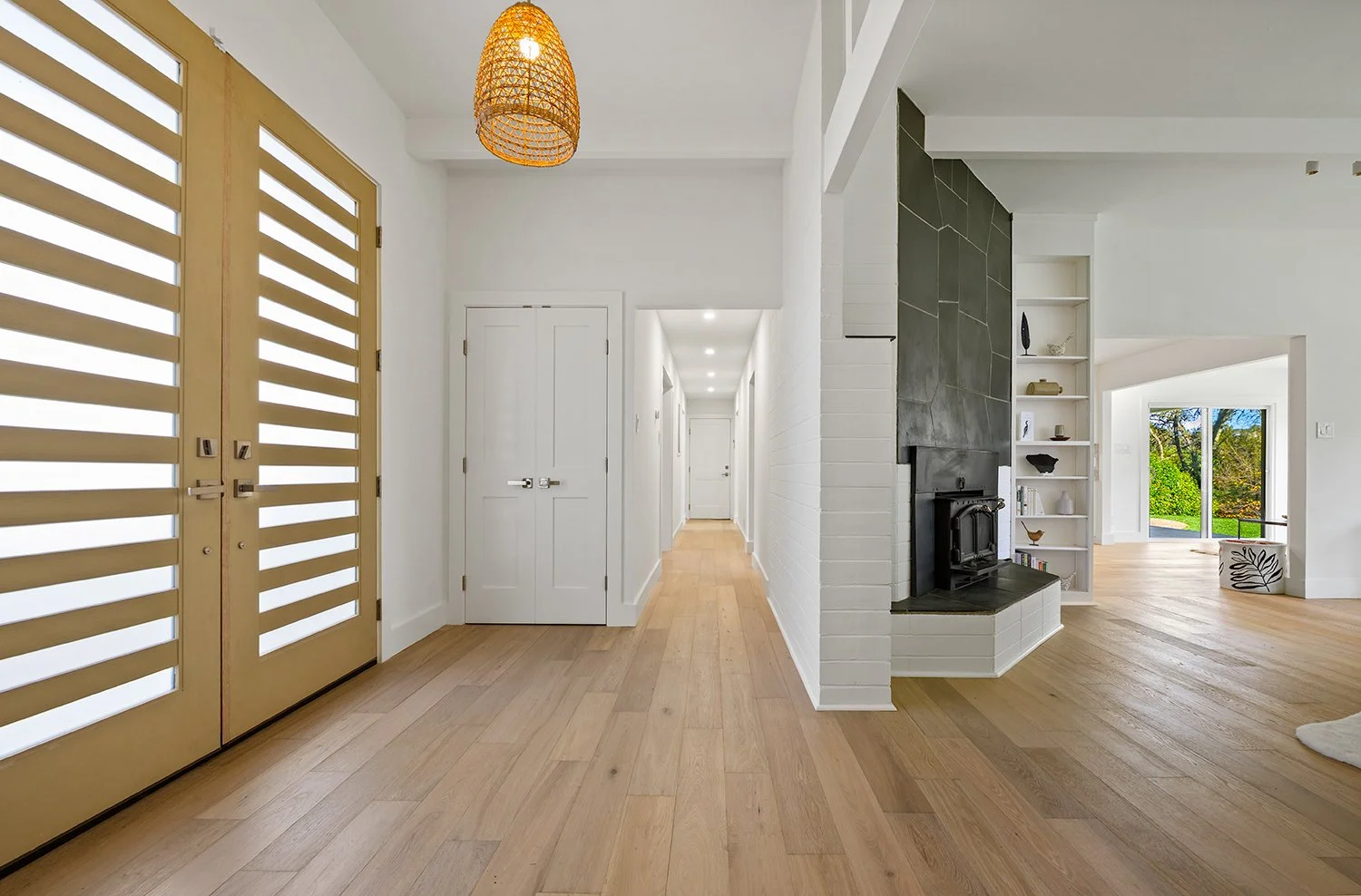 Interior of a modern home with white walls, light wooden floors, a hallway with multiple doors, and a corner with a fireplace and built-in shelves, with sliding glass doors leading to a garden.