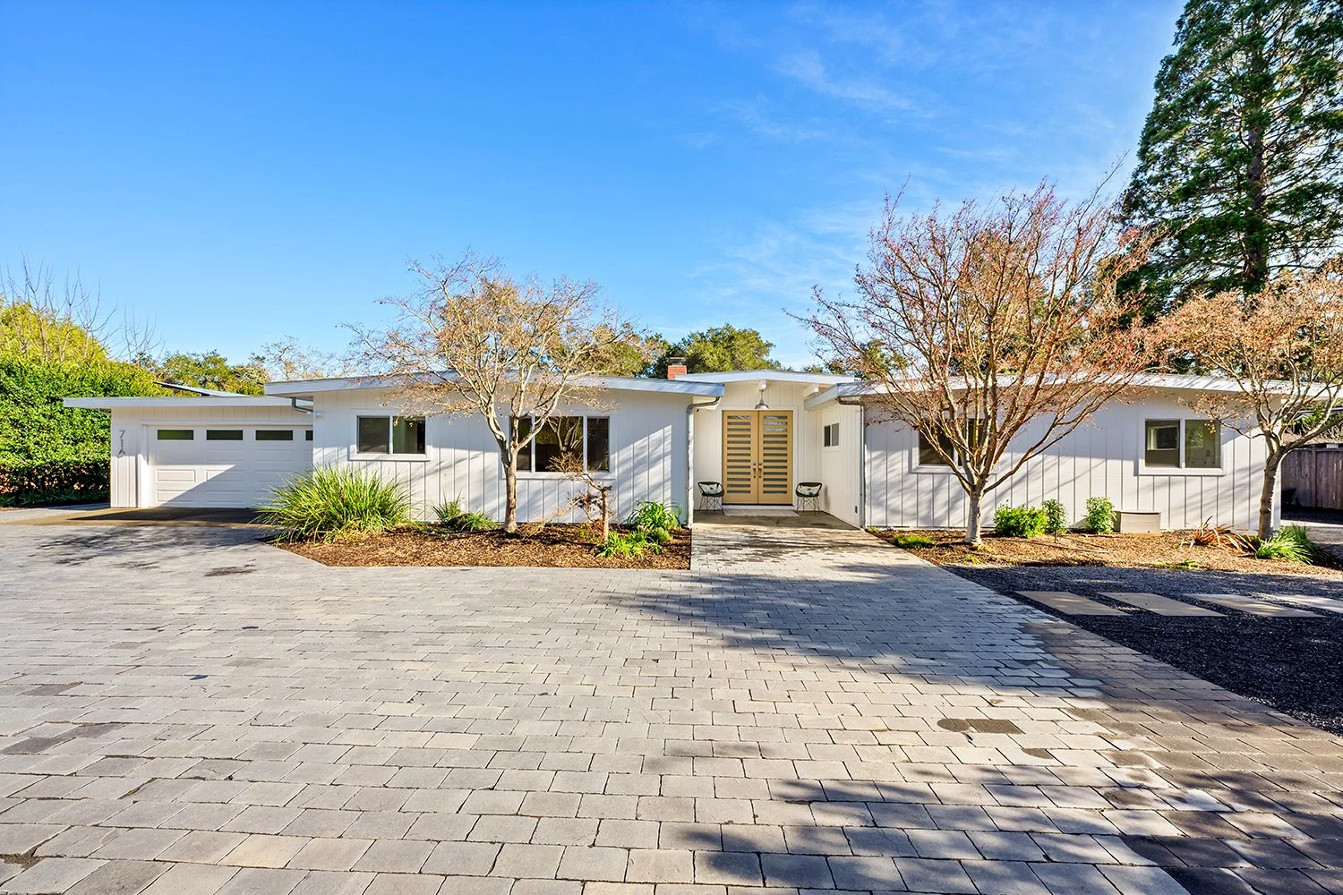 A modern white house with a divided front porch, three trees in the front yard, and a paved driveway leading to a two-car garage, under a clear blue sky.