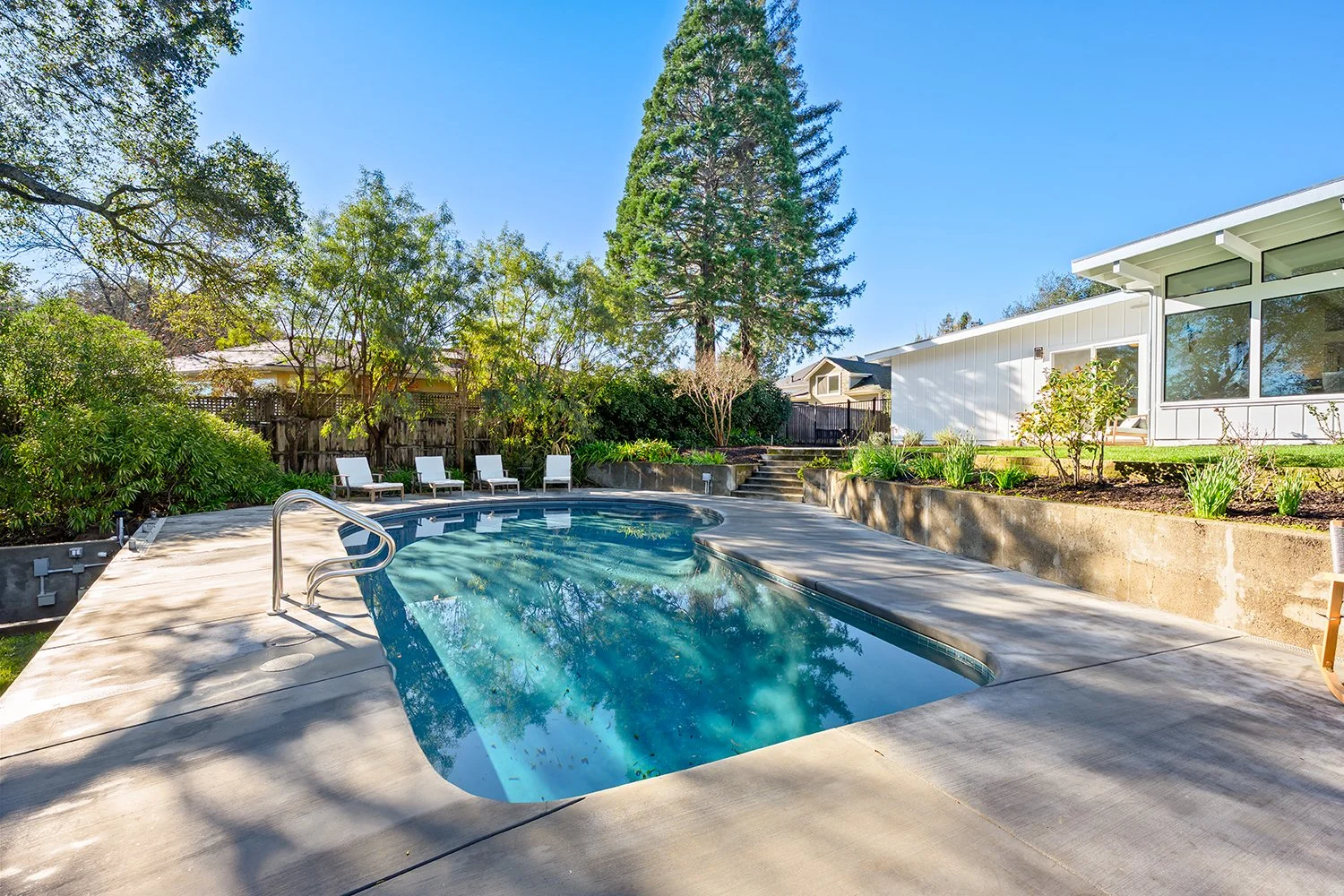 Residential backyard with a kidney-shaped swimming pool, surrounded by trees, shrubs, and several lounge chairs. Modern house with large windows and a garden with plants and flowers.