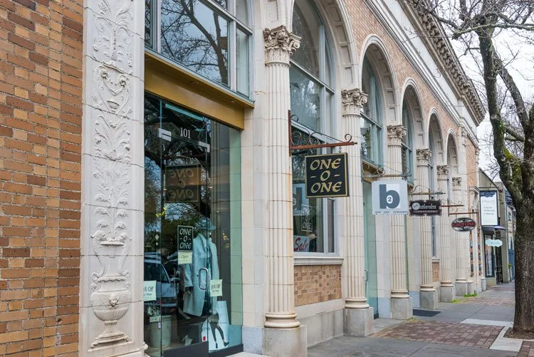Street view of a row of retail stores with ornate architectural details, large windows, and various signs, including one for a store named "ONE of ONE."