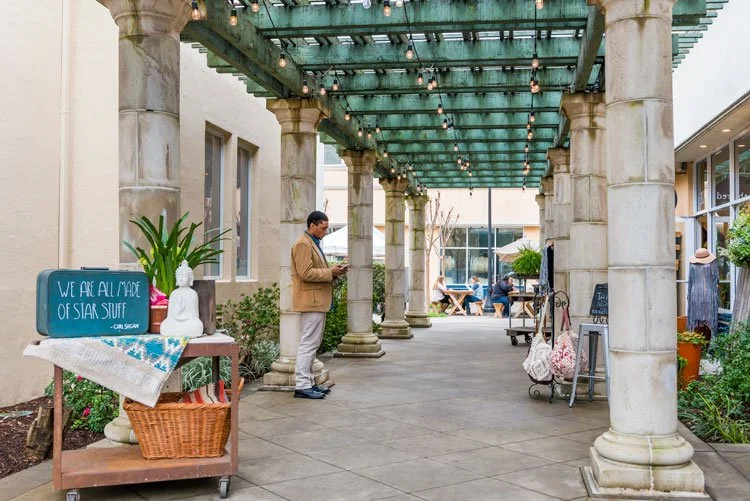 Outdoor shopping or dining area with stone columns supporting a glass-green tiled canopy, with people sitting at tables and a man standing and looking at his phone, surrounded by plants and market stands.