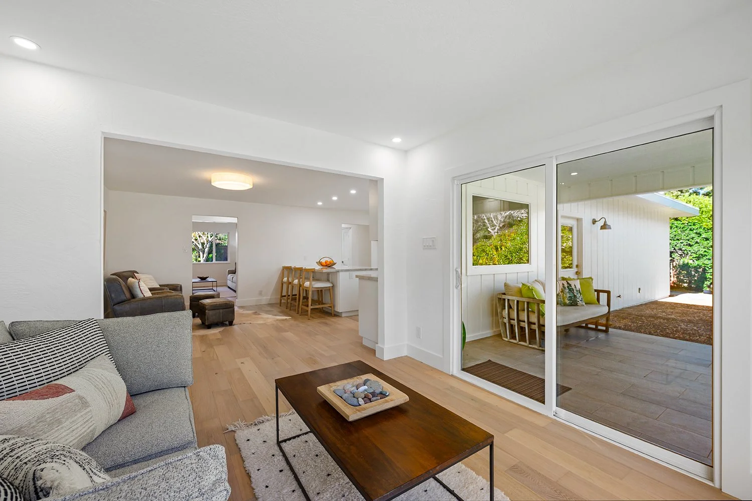 Living room with light-colored walls, hardwood floors, grey sofa with patterned pillows, wooden coffee table with decorative stones, door leading to an outdoor patio with a bench and greenery.